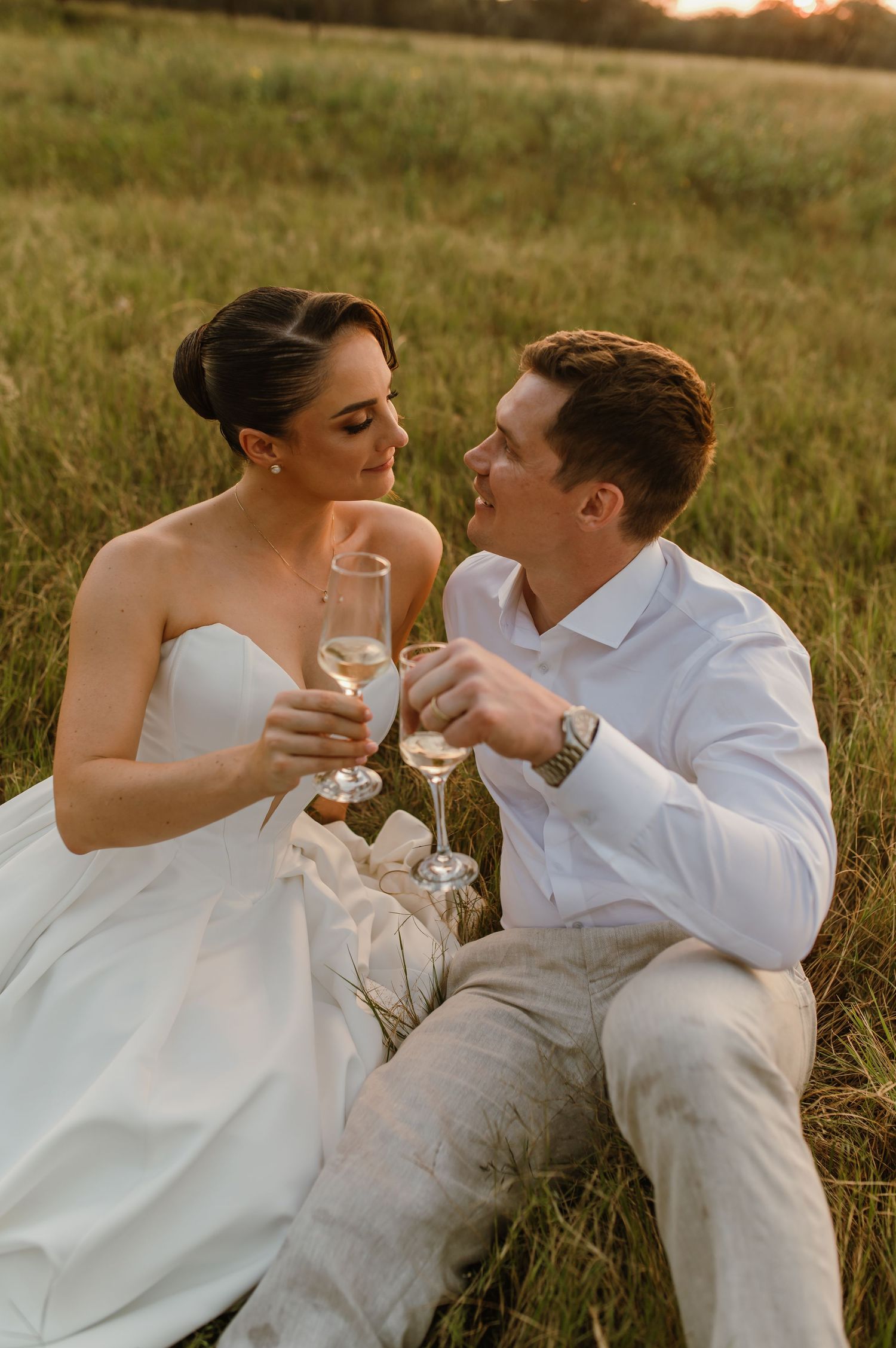 Wedding couple relaxes in tall grass during golden hour while sharing champagne together.