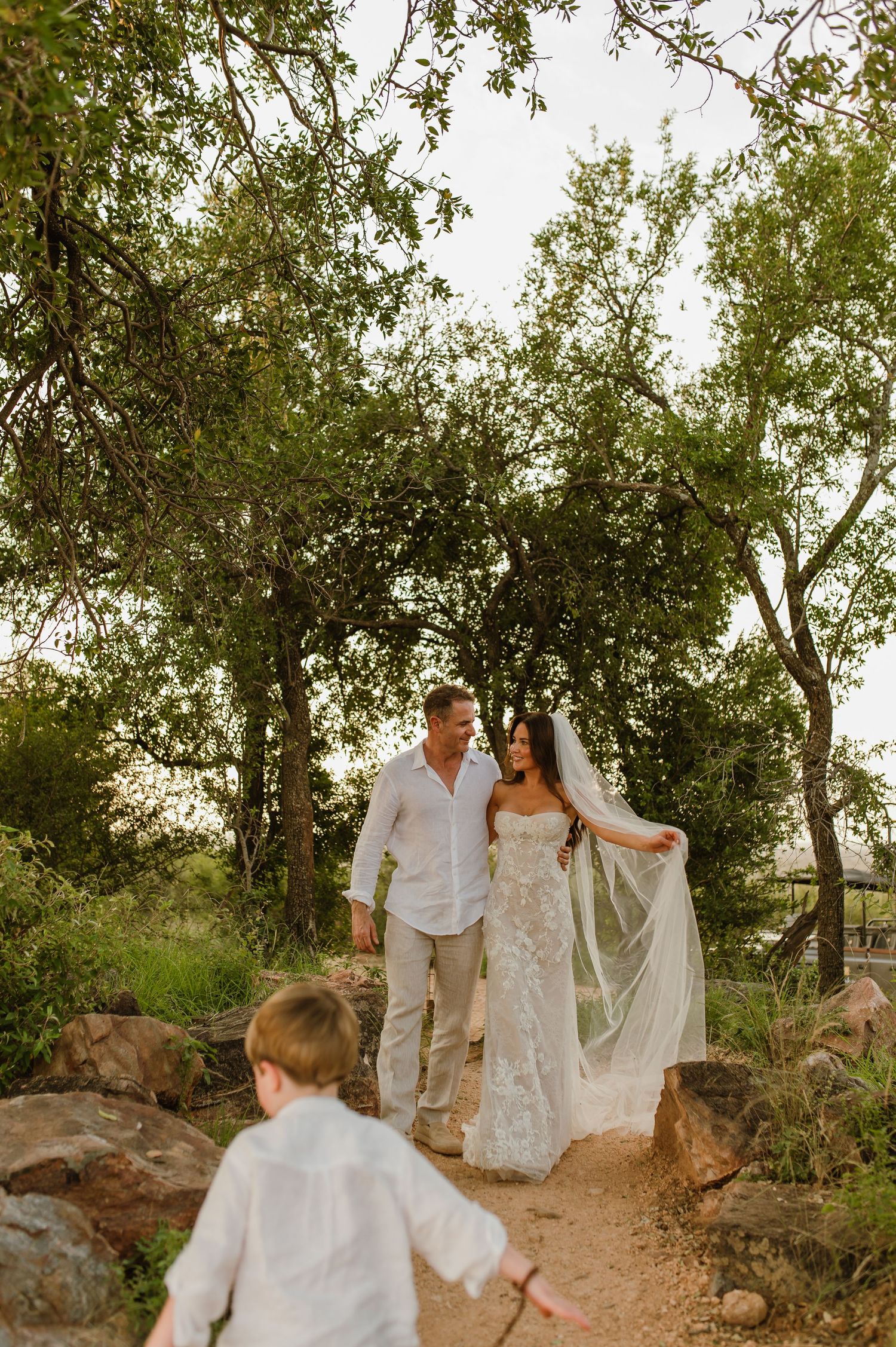 Bride in lace gown and groom walk wooded rocky path as young boy runs ahead during outdoor bush wedding.