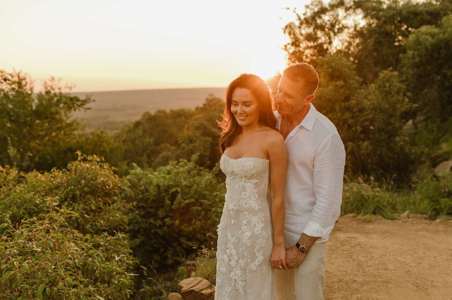 Couple embracing at golden hour sunset on a scenic overlook, bride in strapless lace gown.