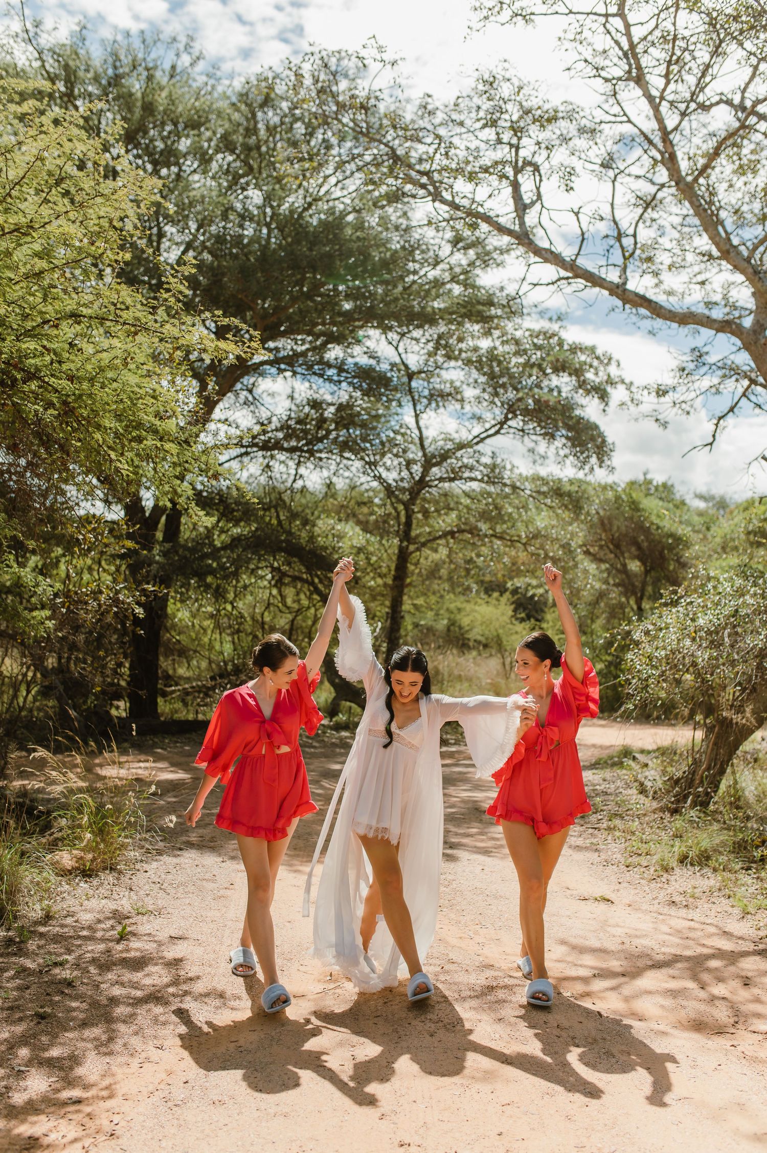 Bride in white gown dances with two bridesmaids in red rompers on a sunny dirt path.