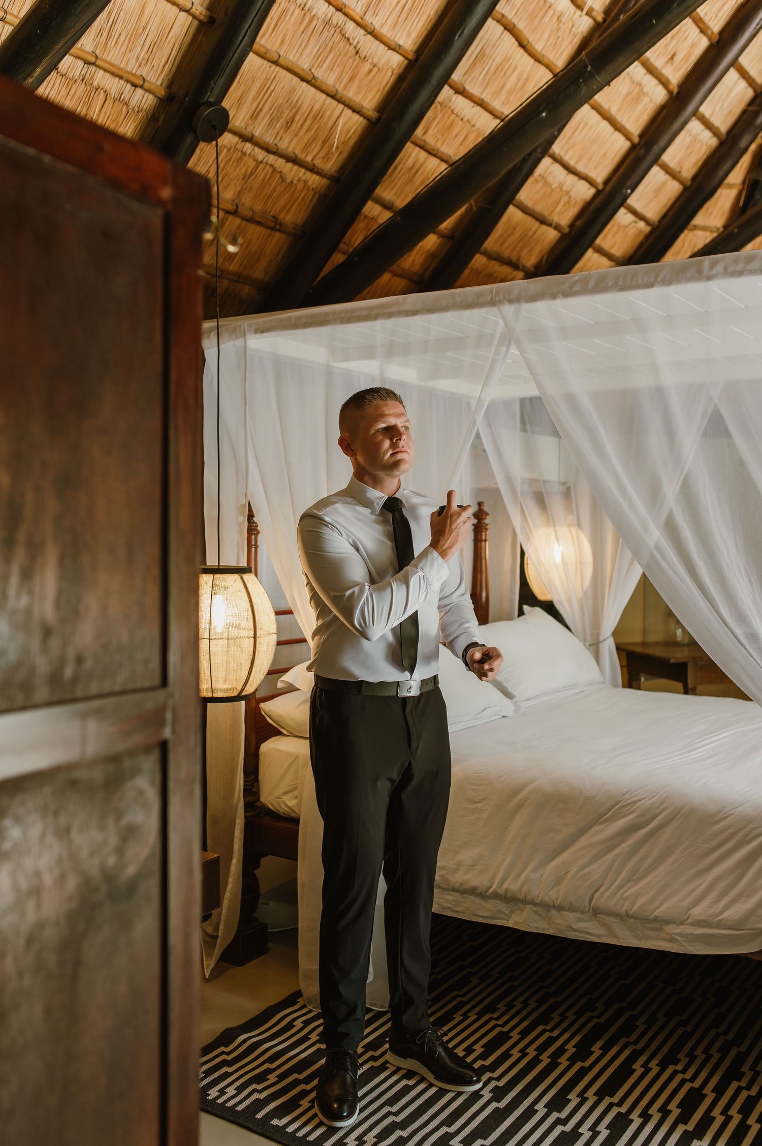 Groom adjusting his tie in a rustic thatched-roof bedroom with a canopy bed before his wedding.