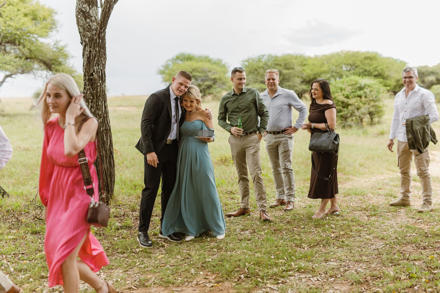 Wedding guests in formal attire gathered outdoors under a tree in a natural African bush setting.