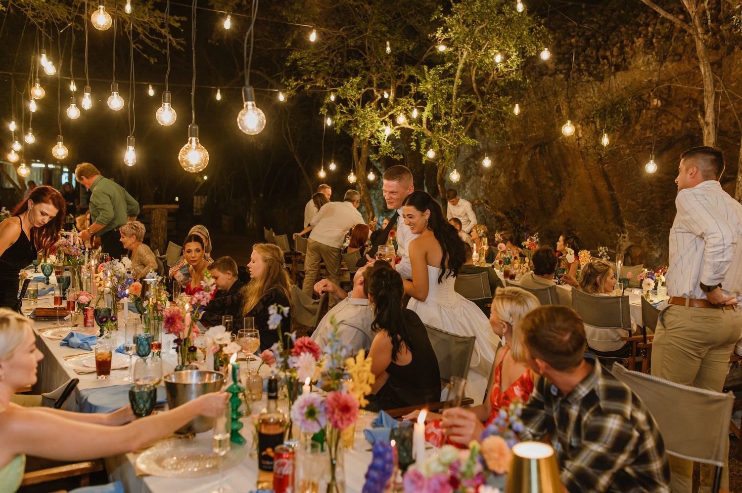 Festive outdoor wedding reception at night with Edison bulb string lights and guests gathered at long decorated tables.