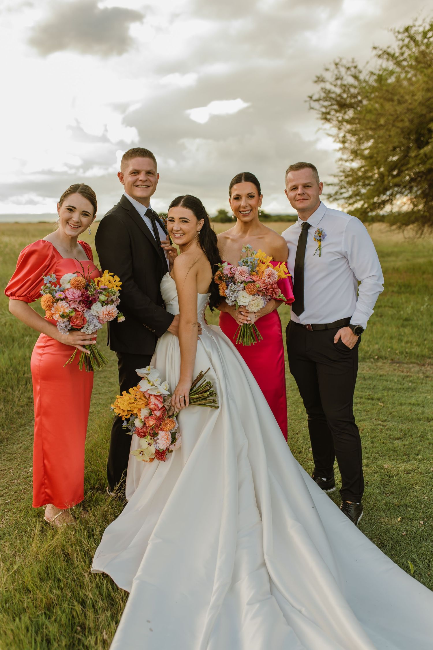 Bride and groom pose with two bridesmaids in coral and pink dresses holding colorful bouquets outdoors.