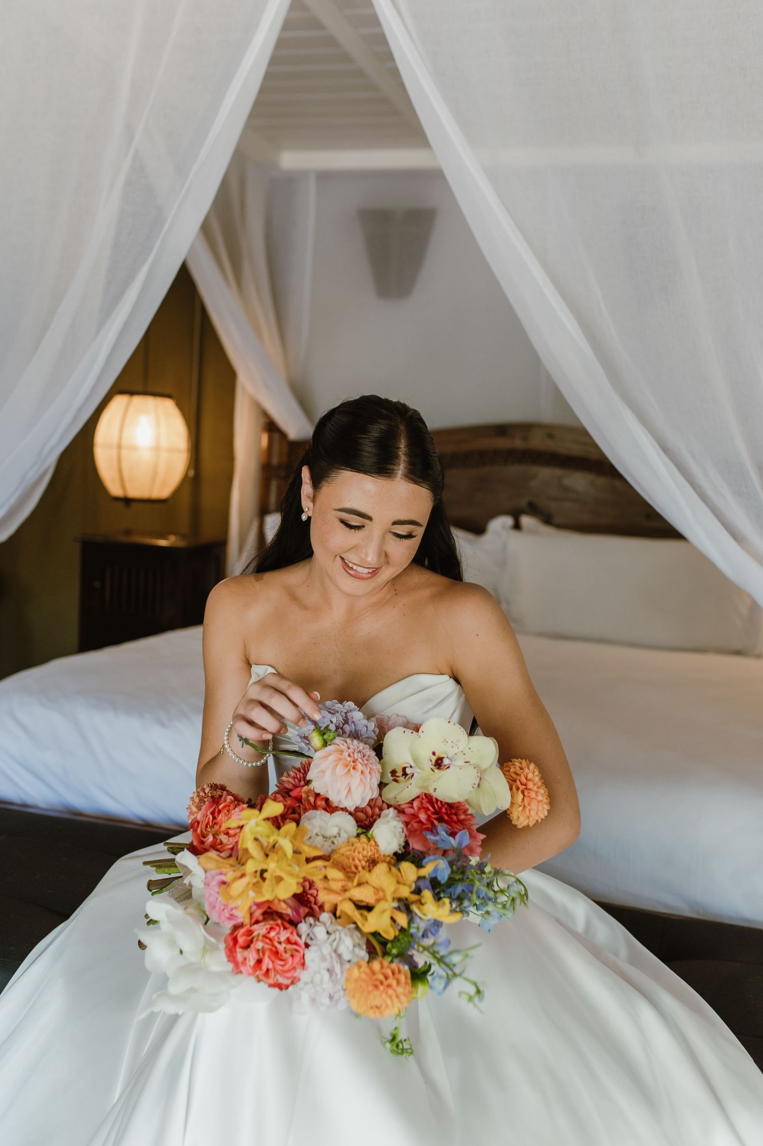 Smiling bride in strapless white gown holding a colorful bouquet of mixed flowers while seated on a canopy bed.