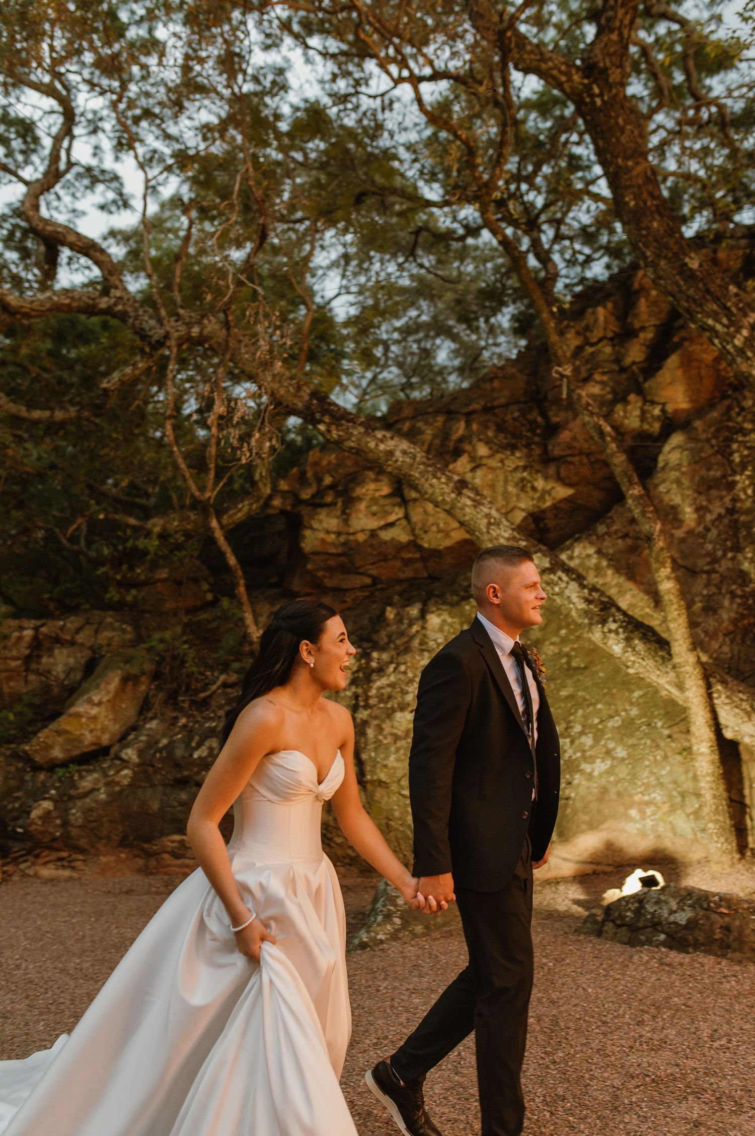Bride and groom walk hand in hand through rustic rocky landscape with warm golden sunset light.