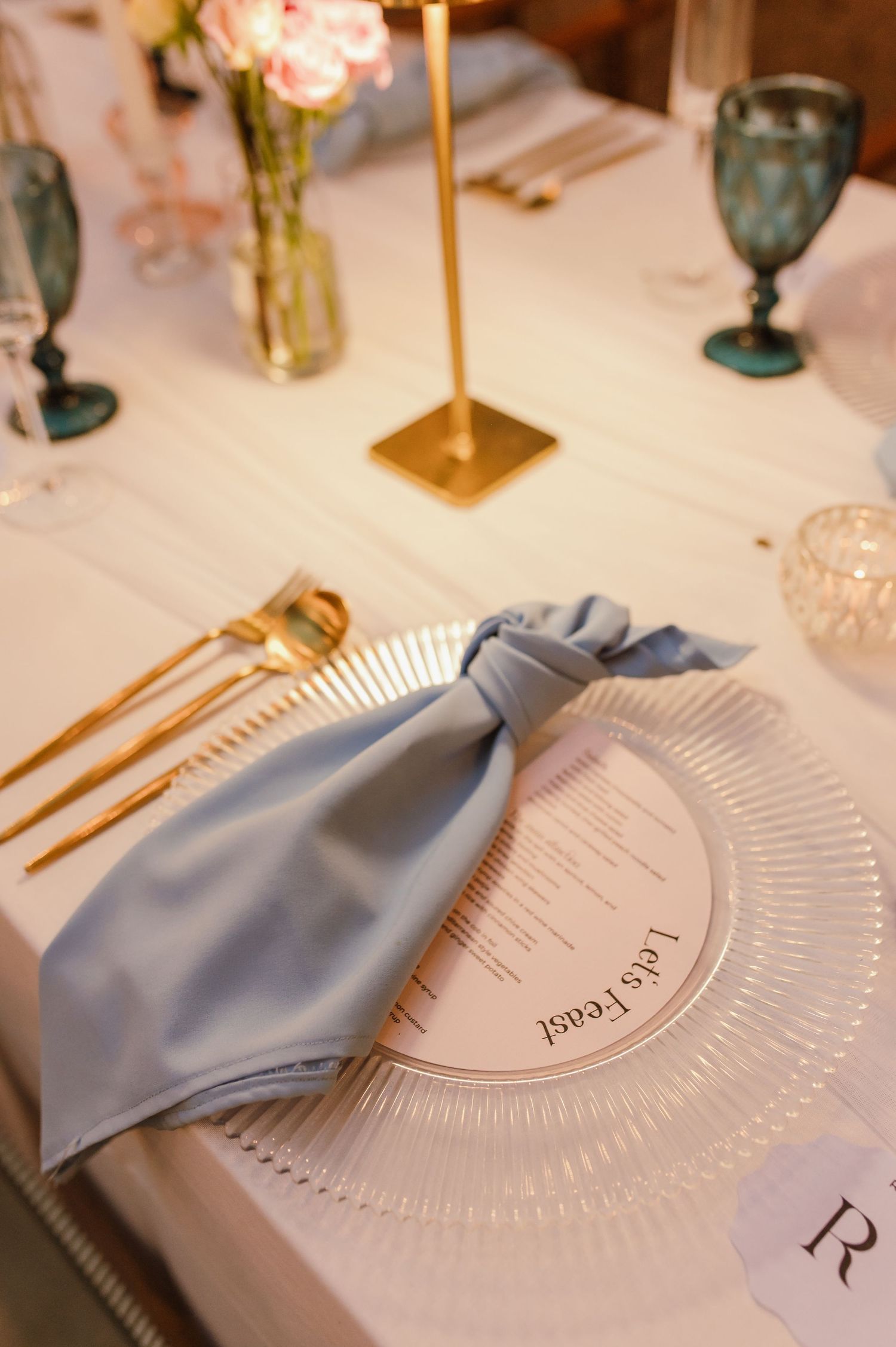 Elegant wedding place setting with gold cutlery, blue silk napkin, circular menu card, and teal glassware on white linen.