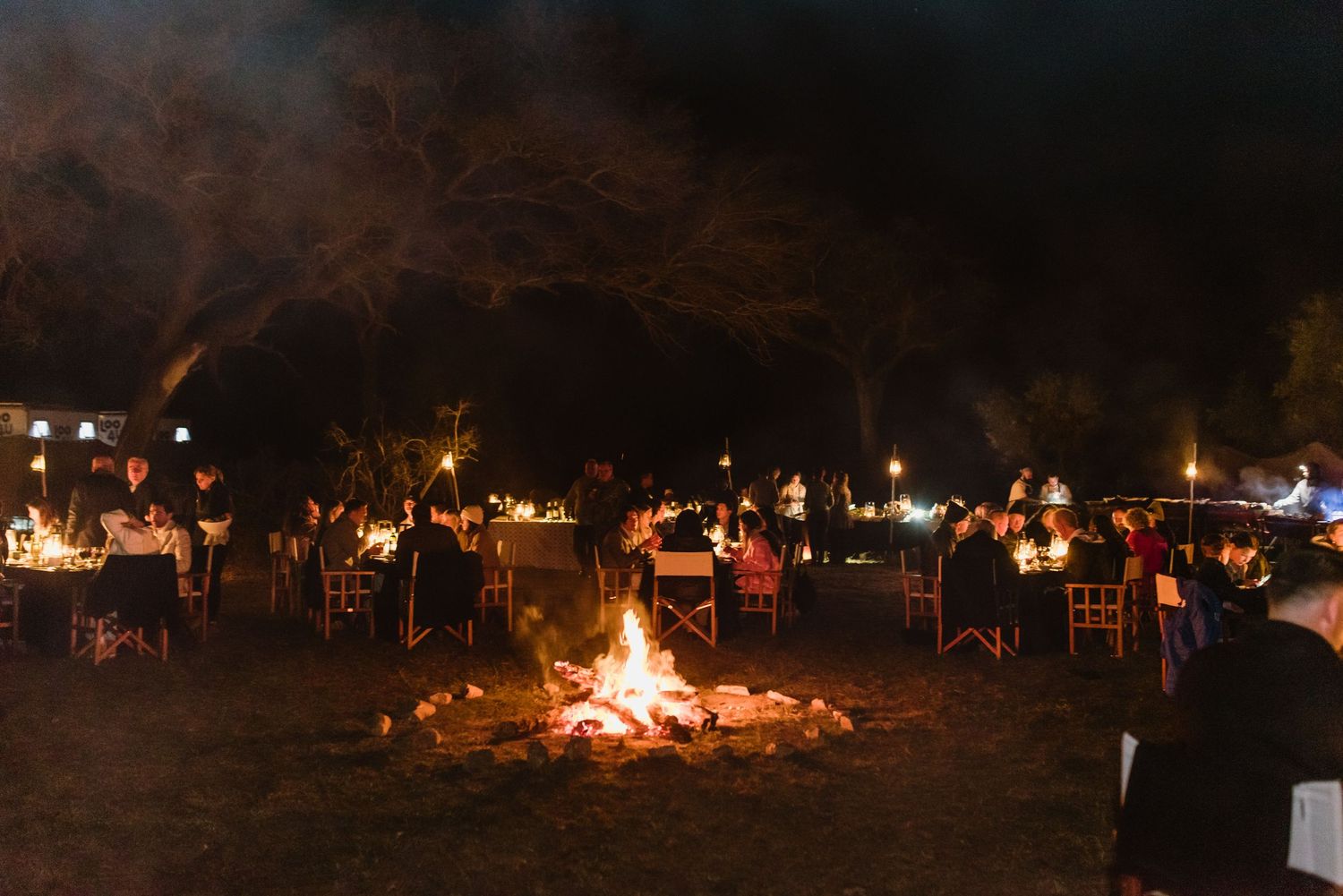 Panoramic view of an outdoor nighttime gathering with multiple bonfires and string lights.