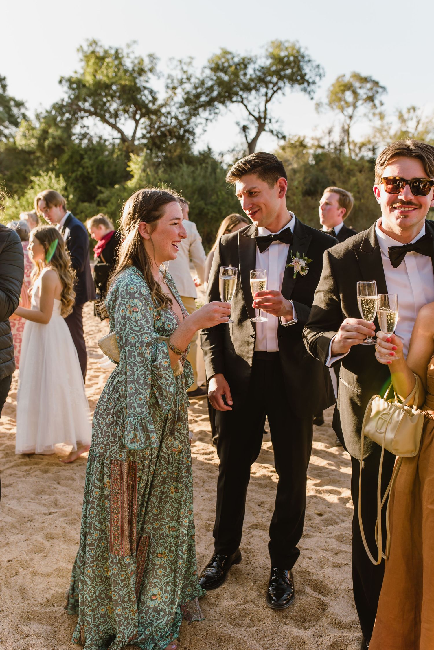 Wedding guests in formal attire mingling and celebrating on a beach during golden hour.