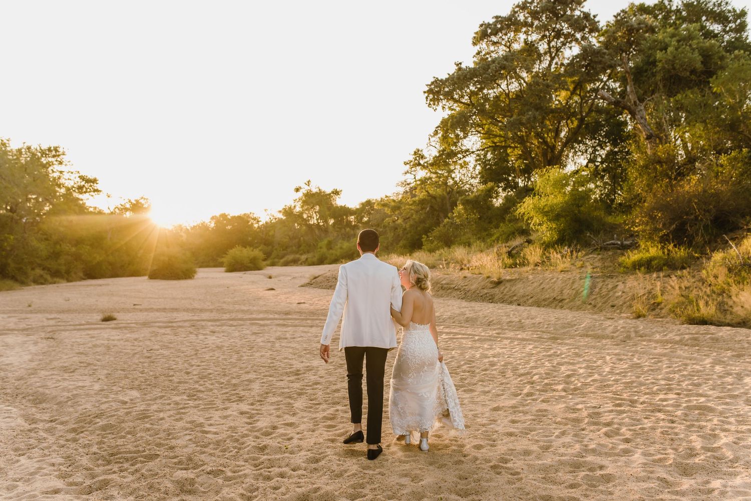 A couple walks hand in hand on a sandy beach at sunset with trees in the background.