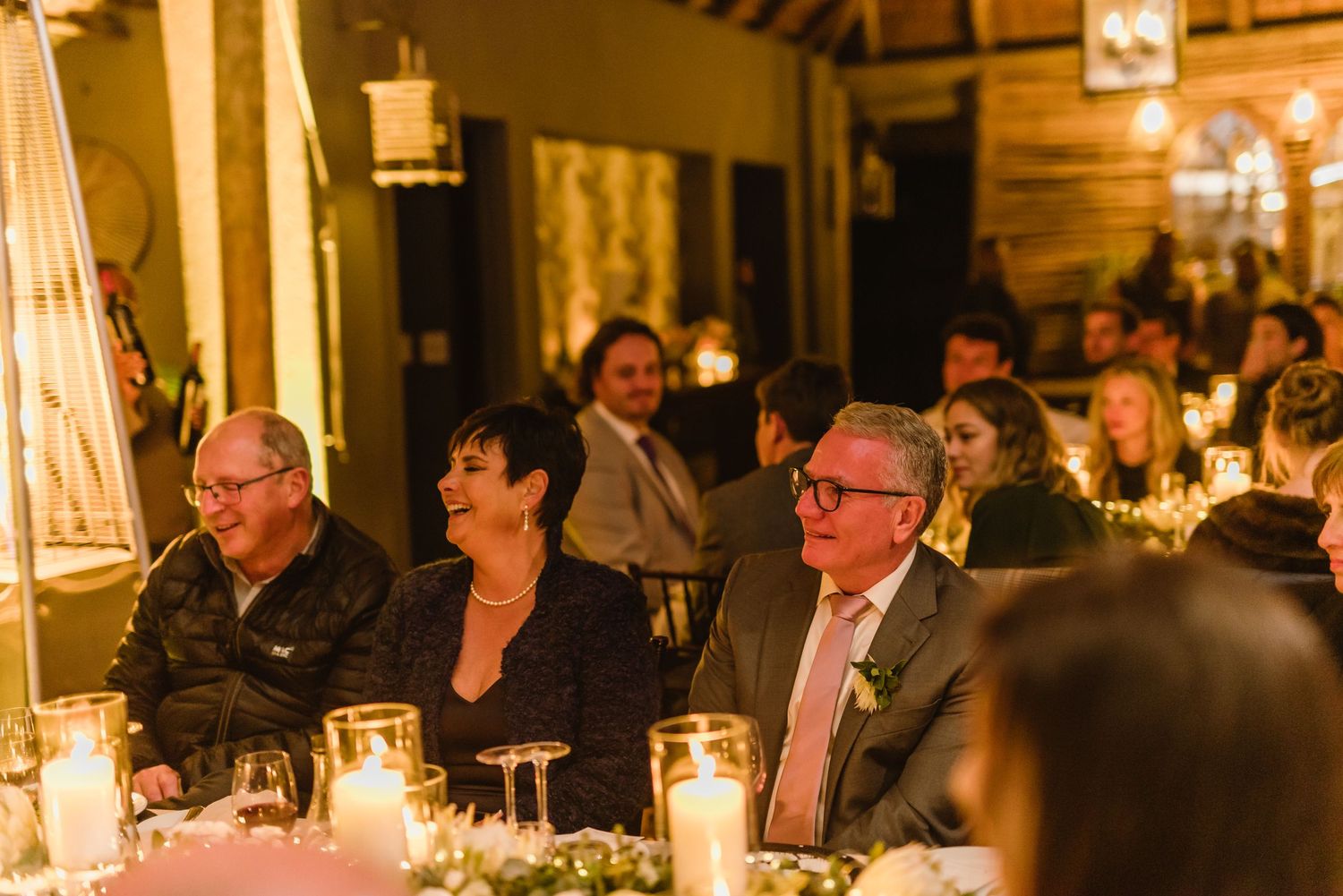 Intimate wedding reception with guests seated at candle-lit tables in a warm, rustic setting.