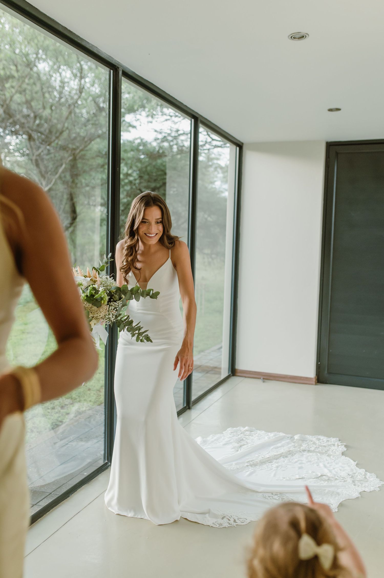 A person in a white wedding gown stands near floor-to-ceiling windows in a modern space.