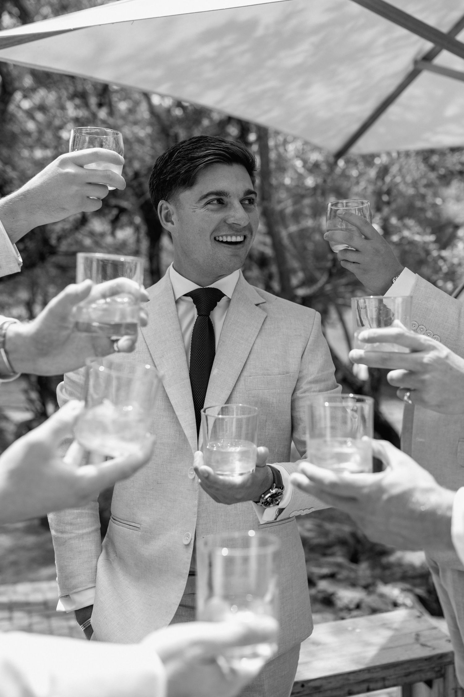 Wedding party in beige suits toasts with champagne glasses under a shaded outdoor setting.