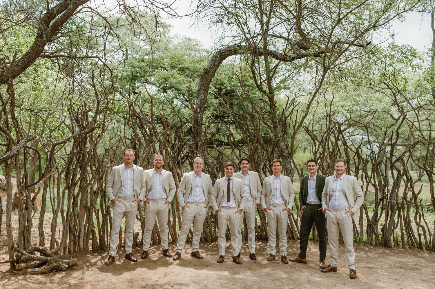 A line of groomsmen in matching suits pose against a backdrop of trees.