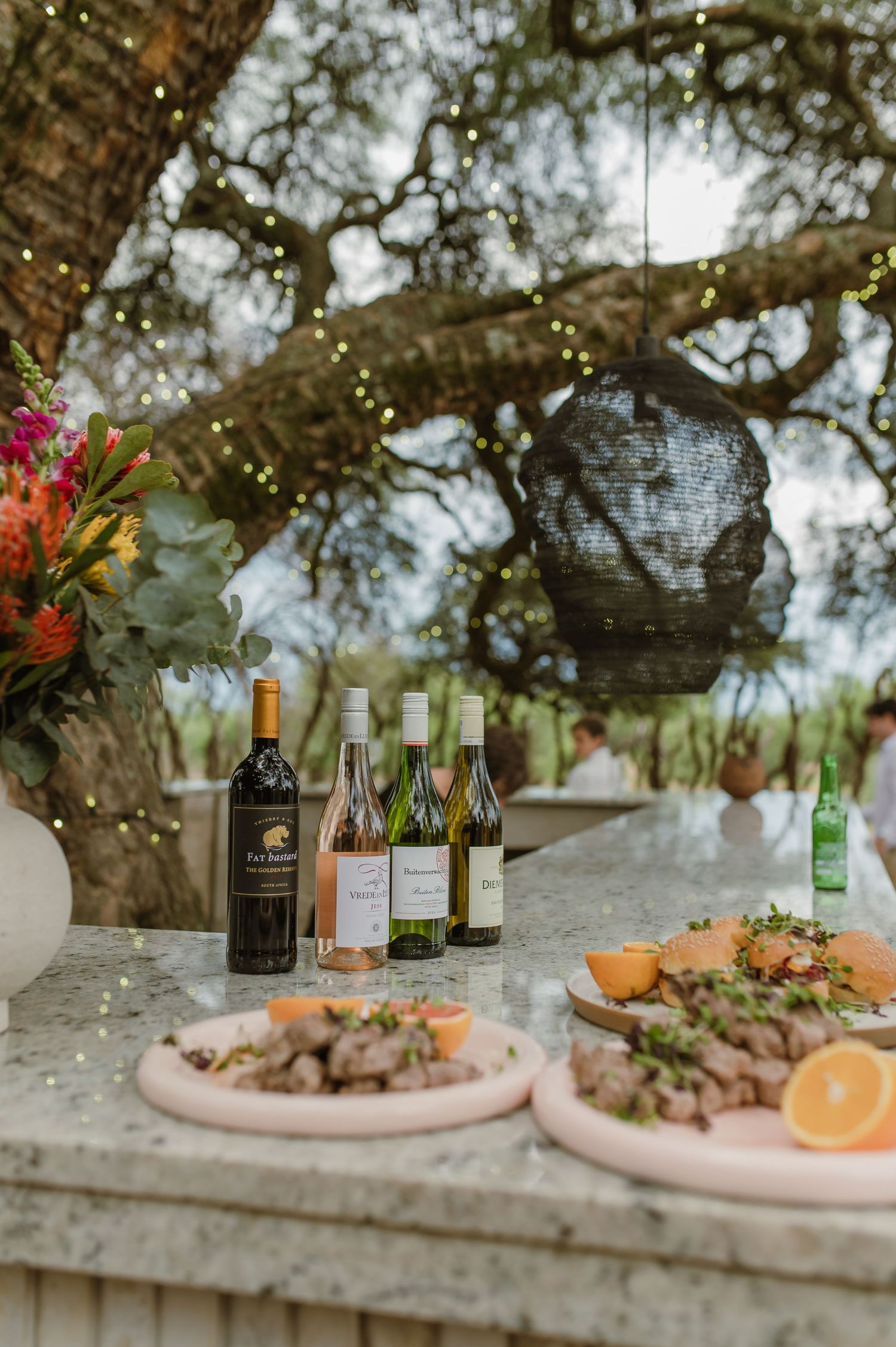 Elegant outdoor wedding reception setup with wine bottles and appetizers on pink plates under a tree.