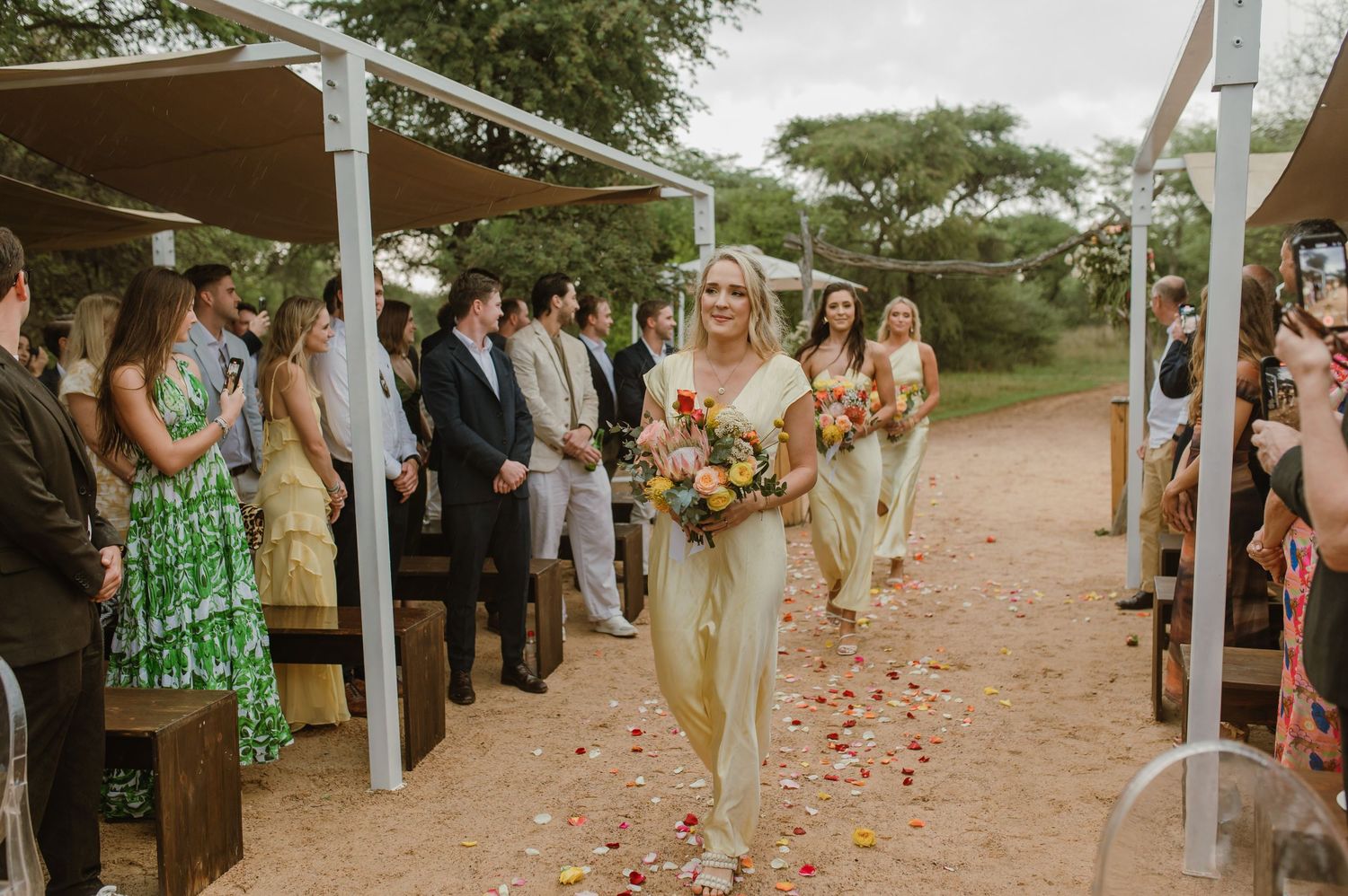 Outdoor wedding ceremony with guests lining a petal-covered aisle under white canopy structures.