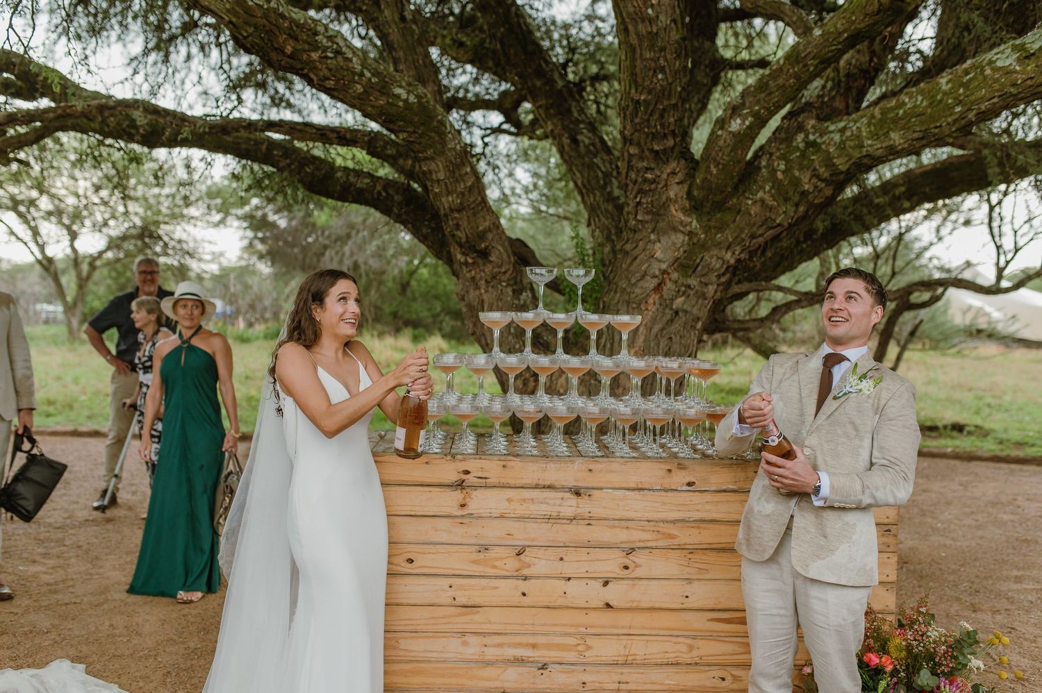 Wedding party gathered under a tree with rustic wooden bar setup and glassware displays.
