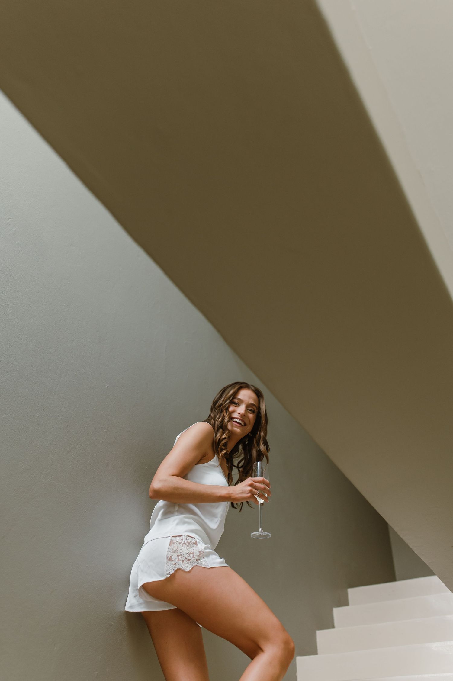 Woman in white lace romper poses with champagne glass on modern staircase.