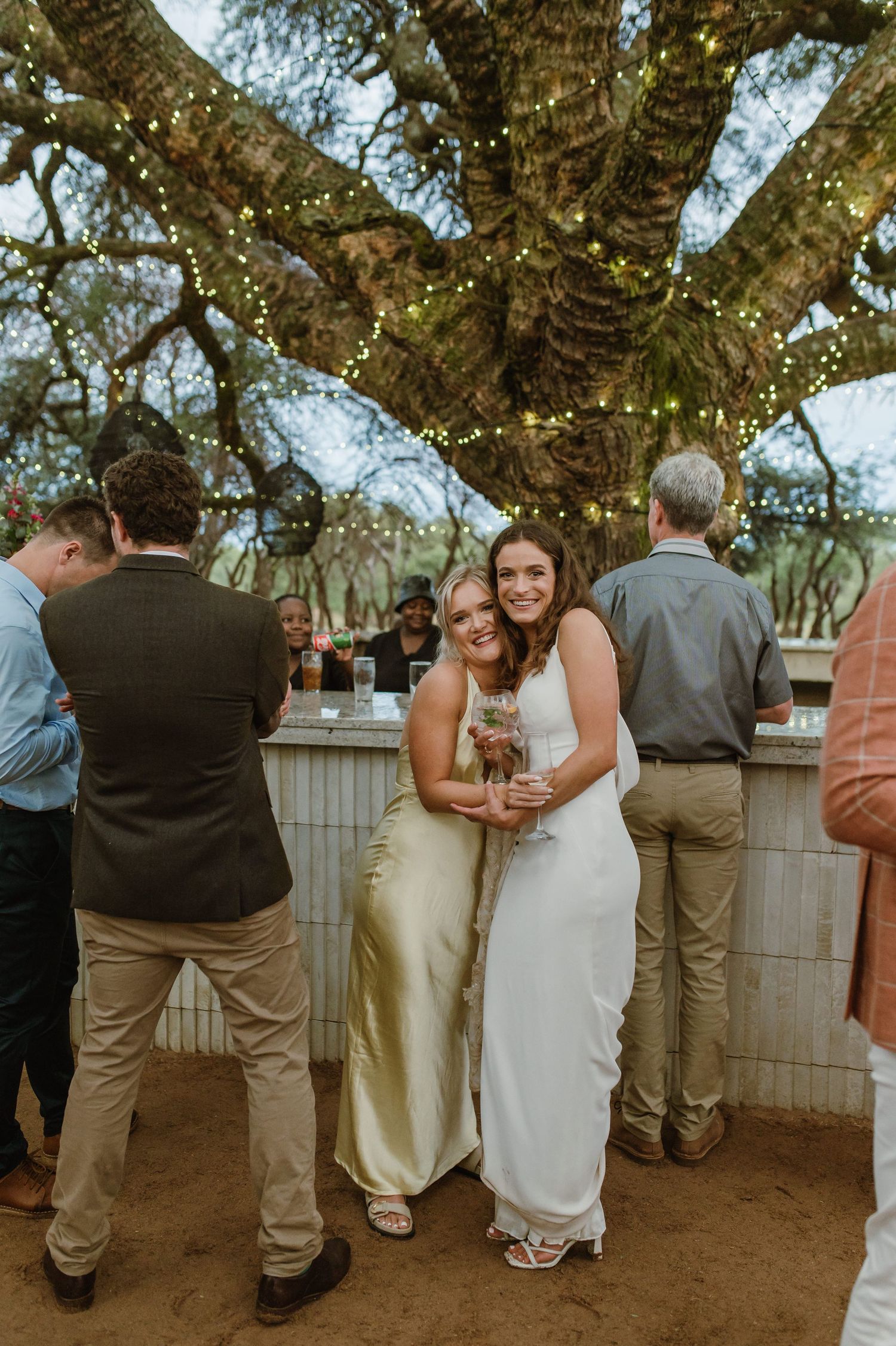 Wedding ceremony guests gather under trees with string lights at an outdoor venue.