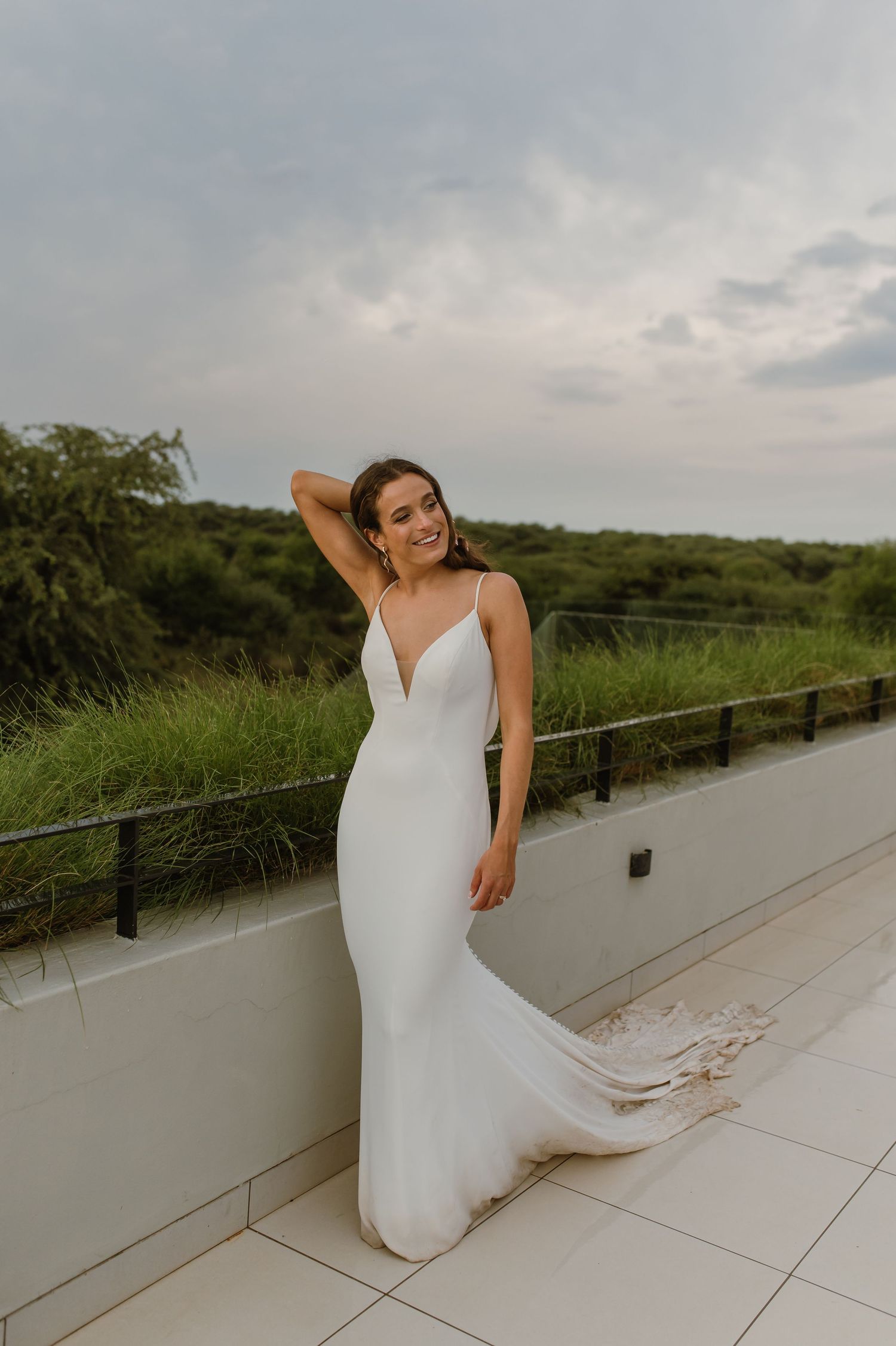 Bride in sleek white wedding gown poses against natural landscape.