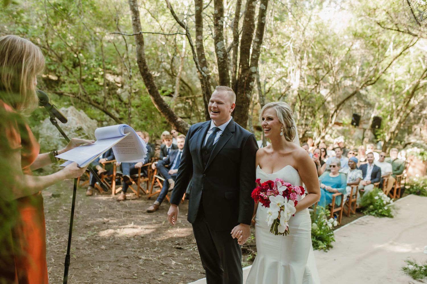 Outdoor forest wedding ceremony with guests gathered beneath tall trees on a sunny day.