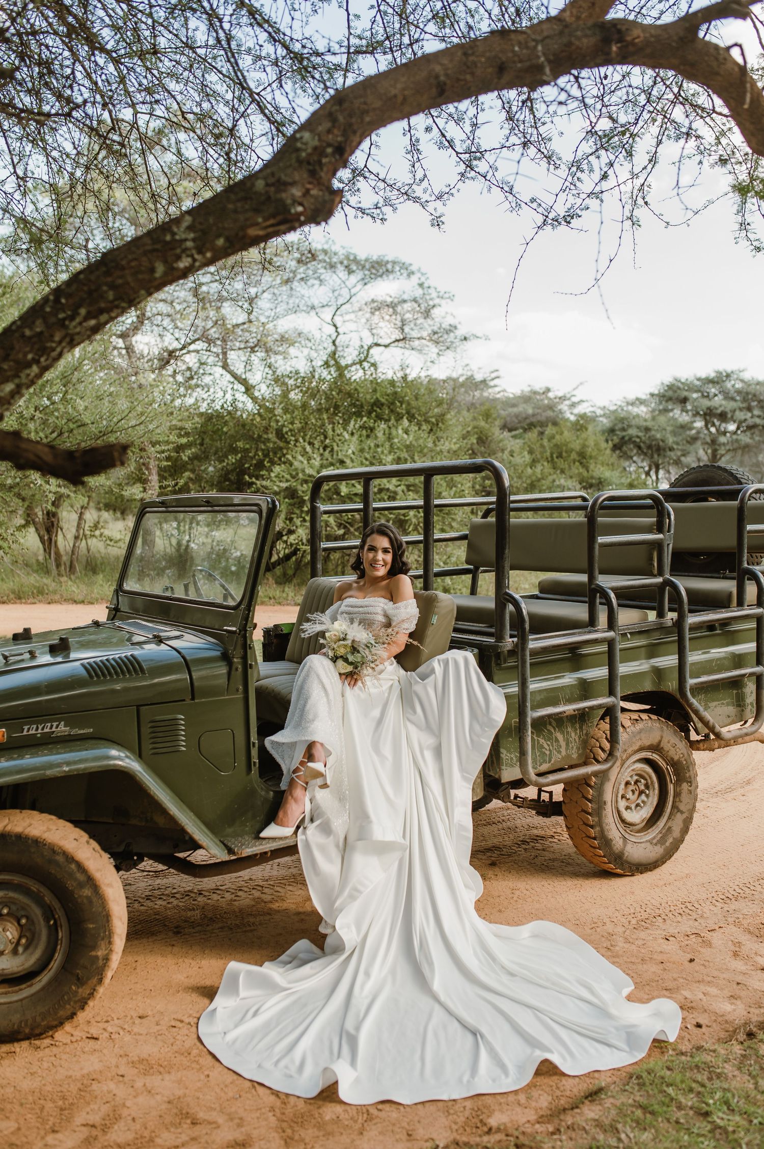 White wedding dress photographed near vintage military vehicles under acacia trees in an outdoor setting.