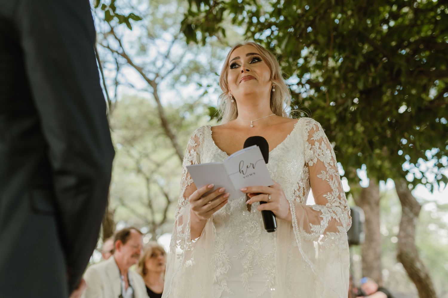 A bride in a lace wedding dress reads from a paper during an outdoor ceremony under trees.