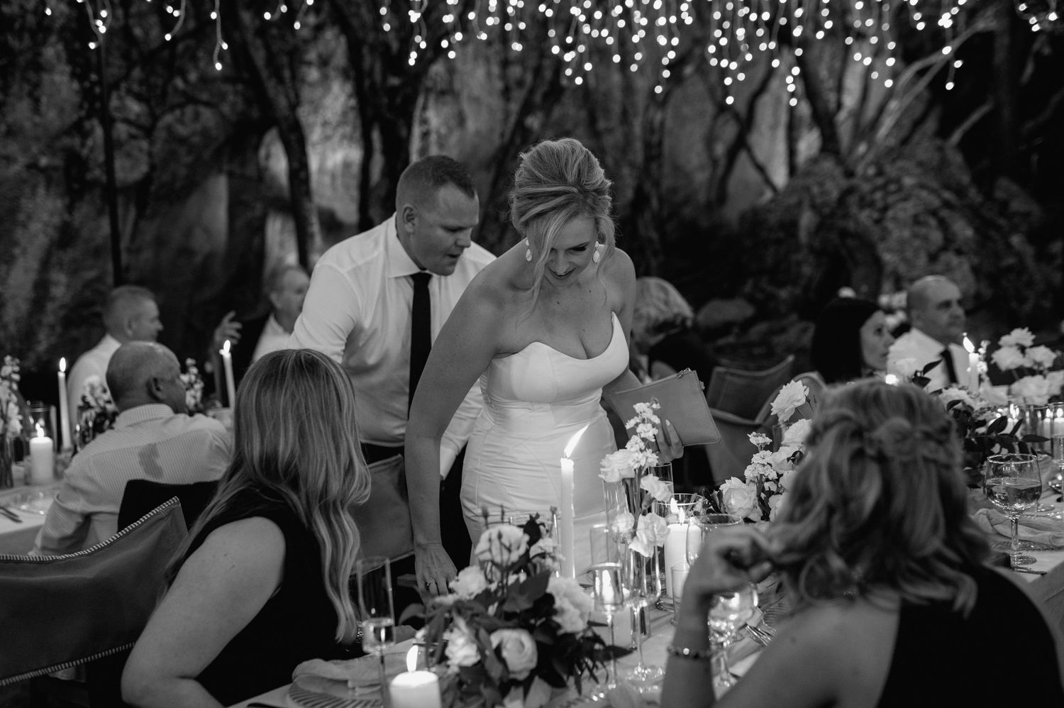 Black and white photo of wedding guests dining under romantic string light decorations.