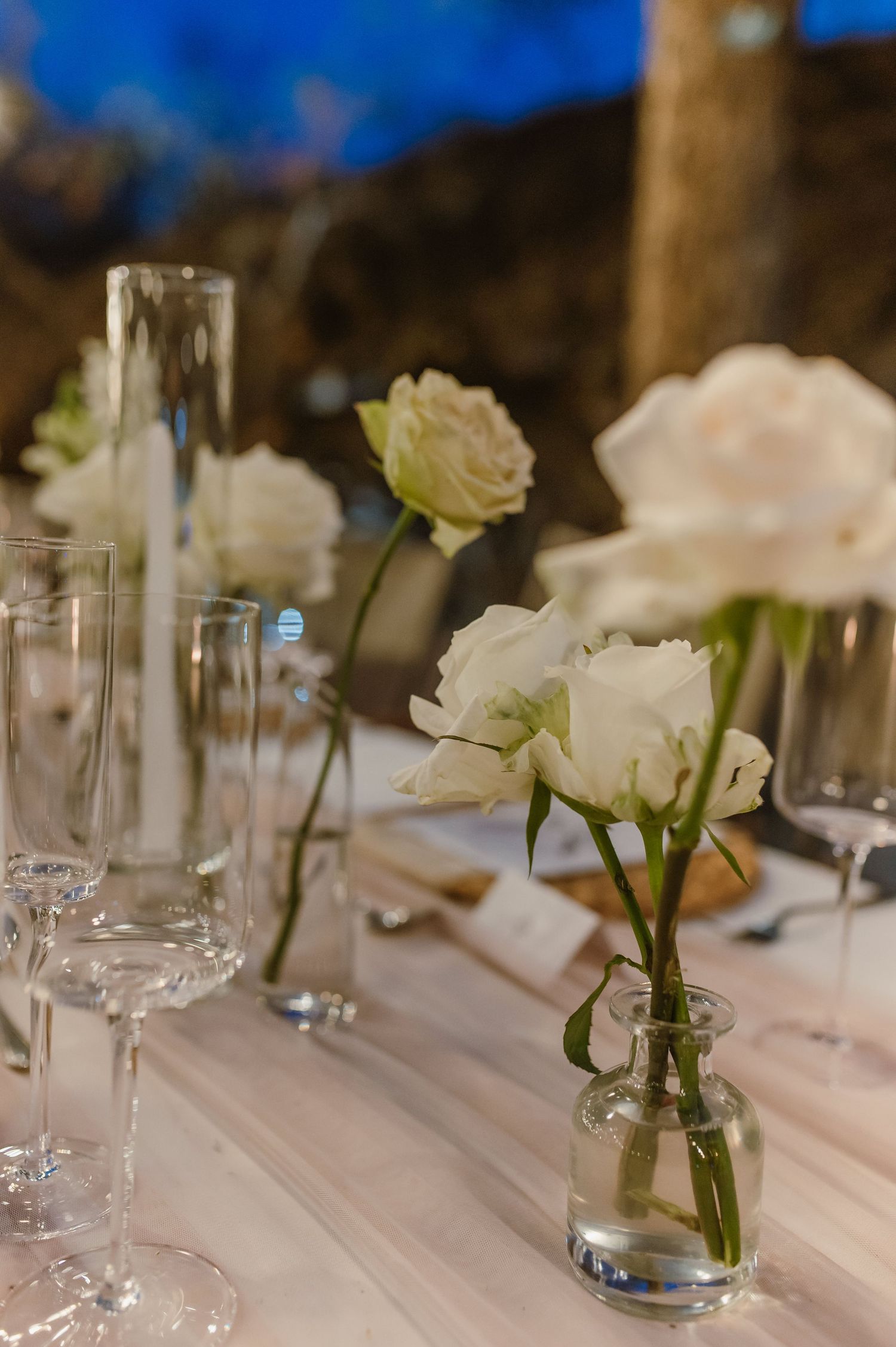 White floral centerpieces in glass vases line a formal dining table with crystal glassware.