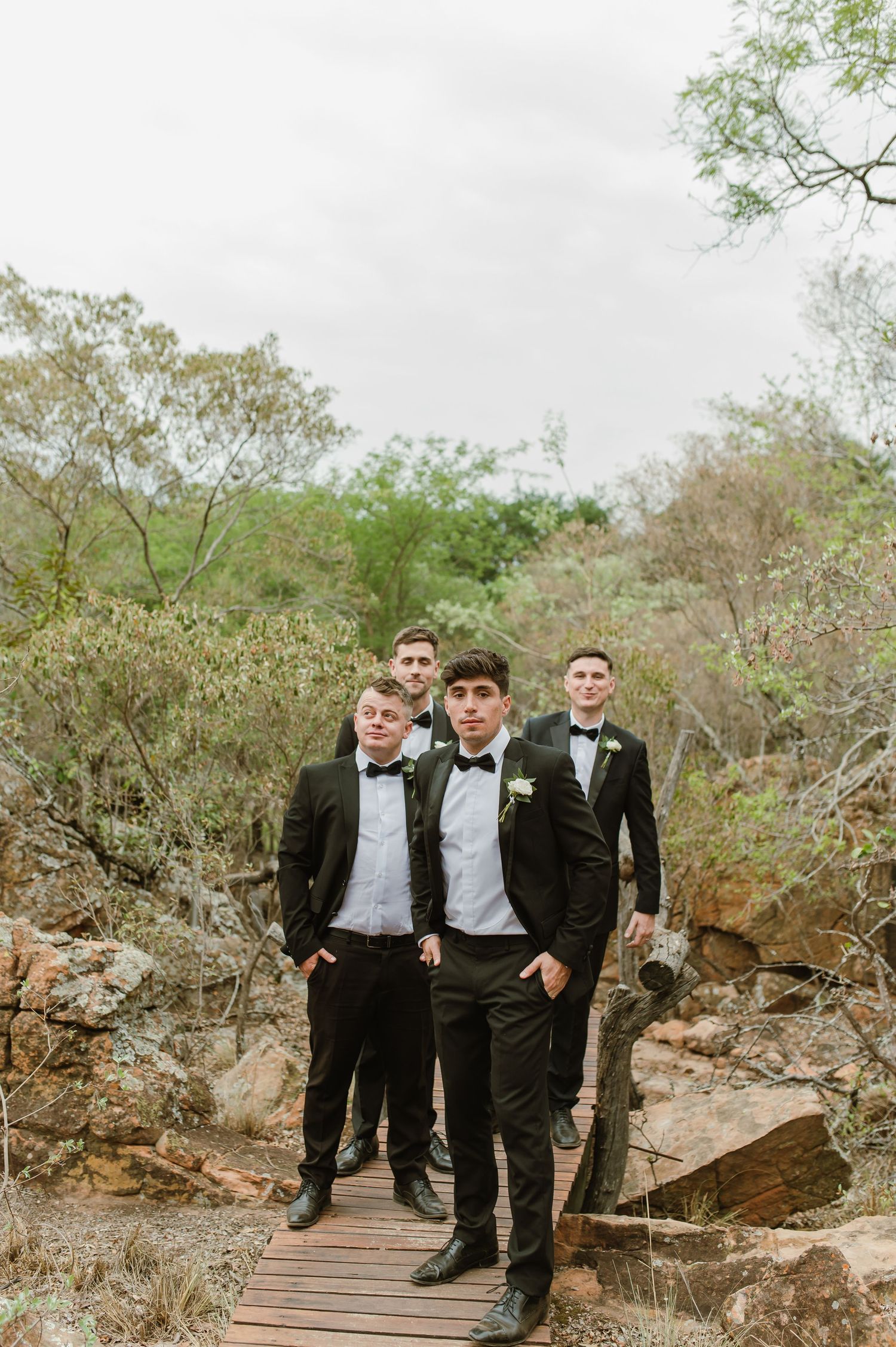 Wedding party members in formal attire gather on a wooden walkway amidst natural rocky terrain.