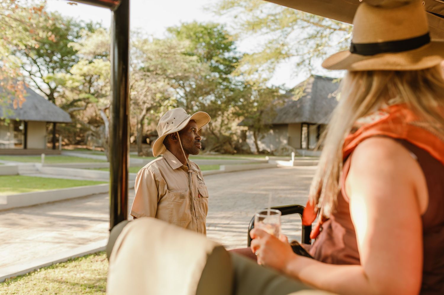 Two travelers enjoy refreshments at an outdoor safari lodge surrounded by thatched-roof buildings.