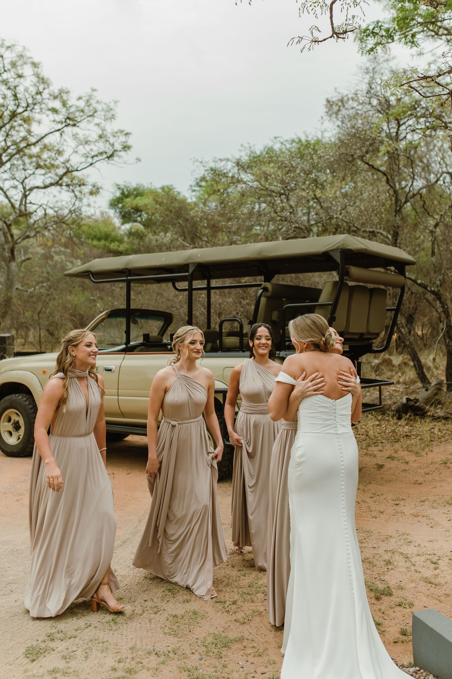 Wedding party in neutral dresses shares joyful moment by safari vehicle in natural setting.