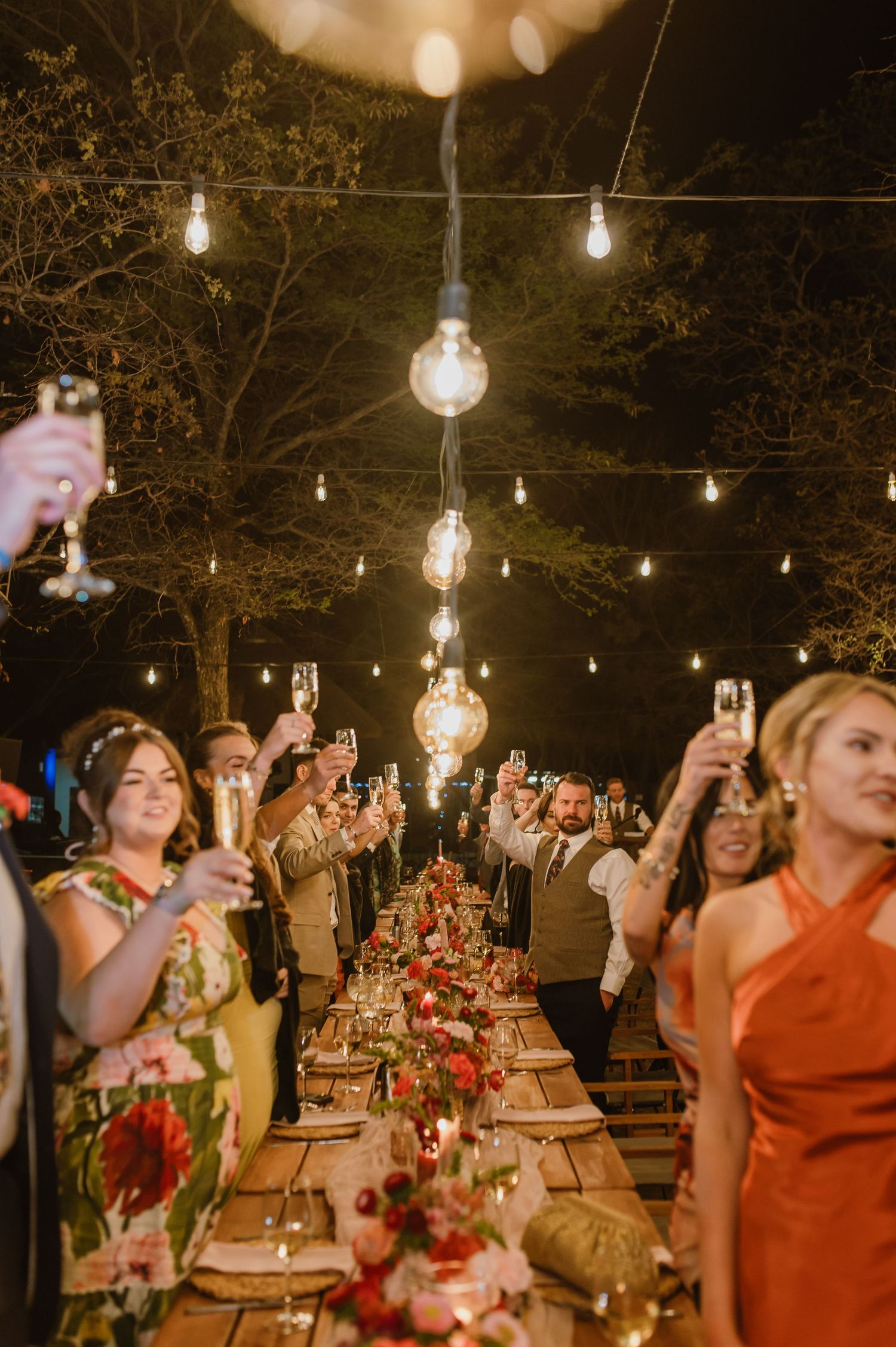 Wedding reception celebration with guests raising glasses under string lights and floral decorations.