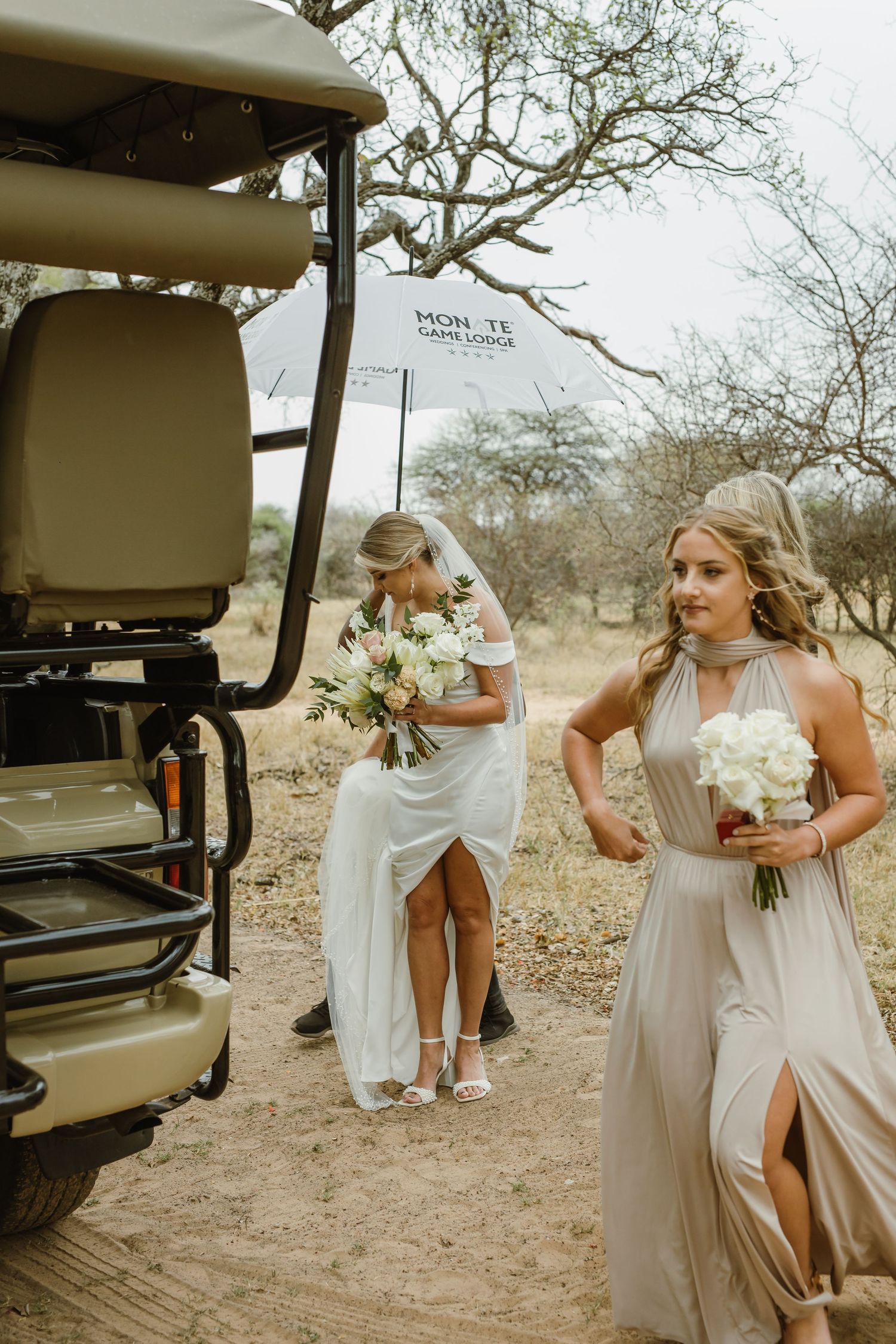Bridesmaids in champagne dresses gather near safari vehicles in a desert setting.