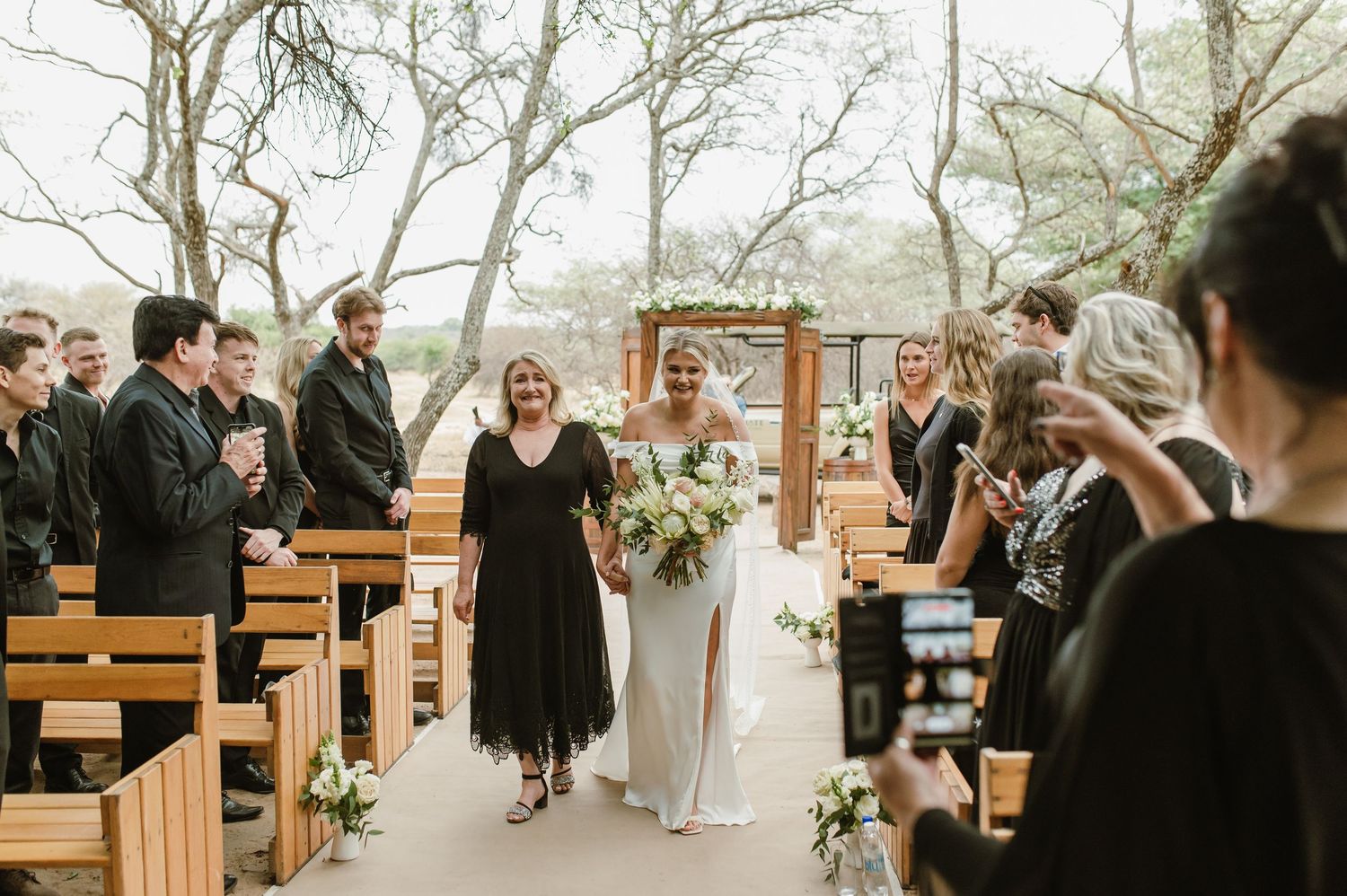 Wedding guests seated on wooden benches during an outdoor ceremony under trees.