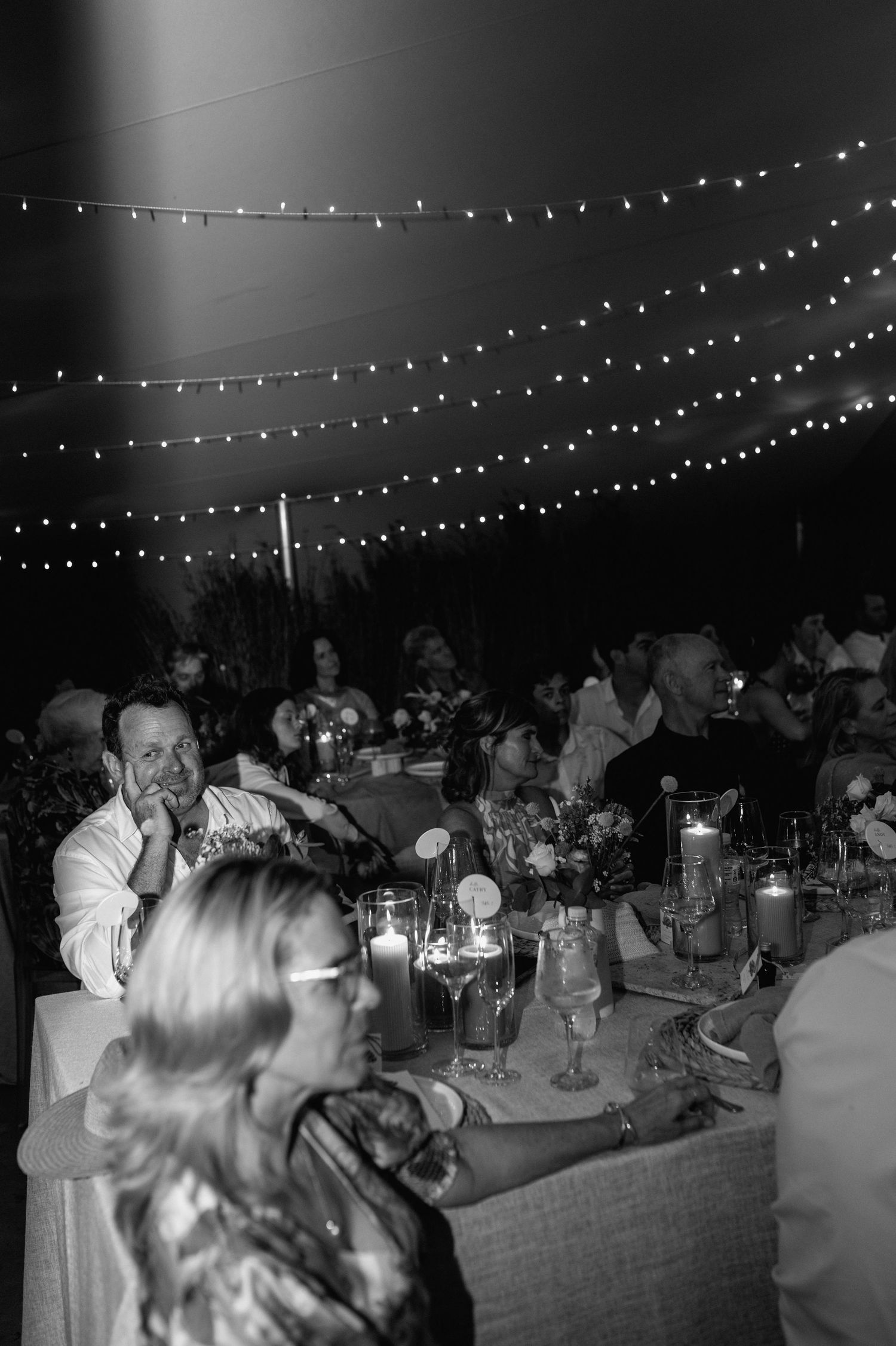 Wedding reception guests seated at long tables under romantic string lights at an evening outdoor celebration.