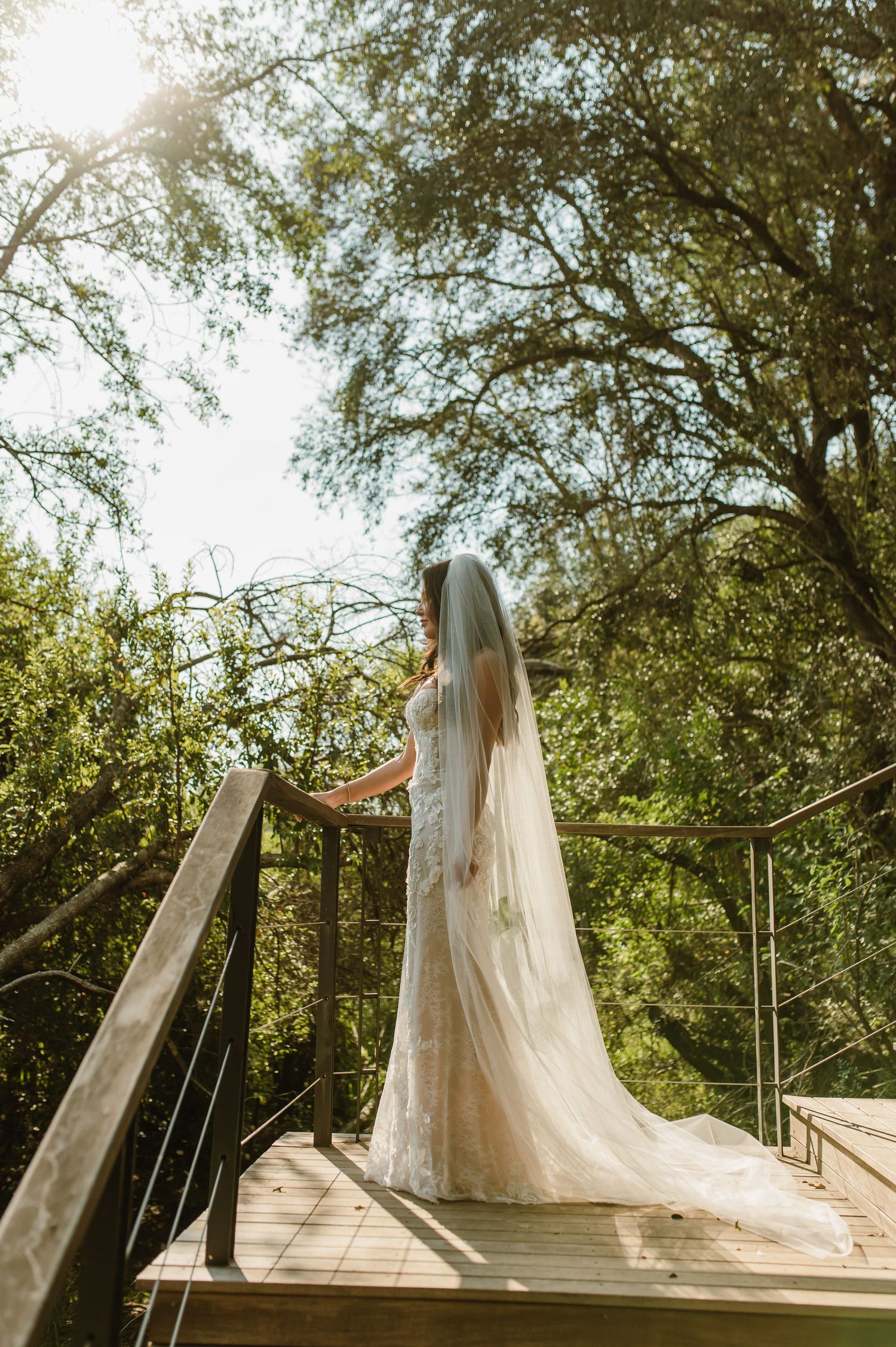 Bride in lace wedding gown and veil standing on treehouse deck surrounded by tall green trees.