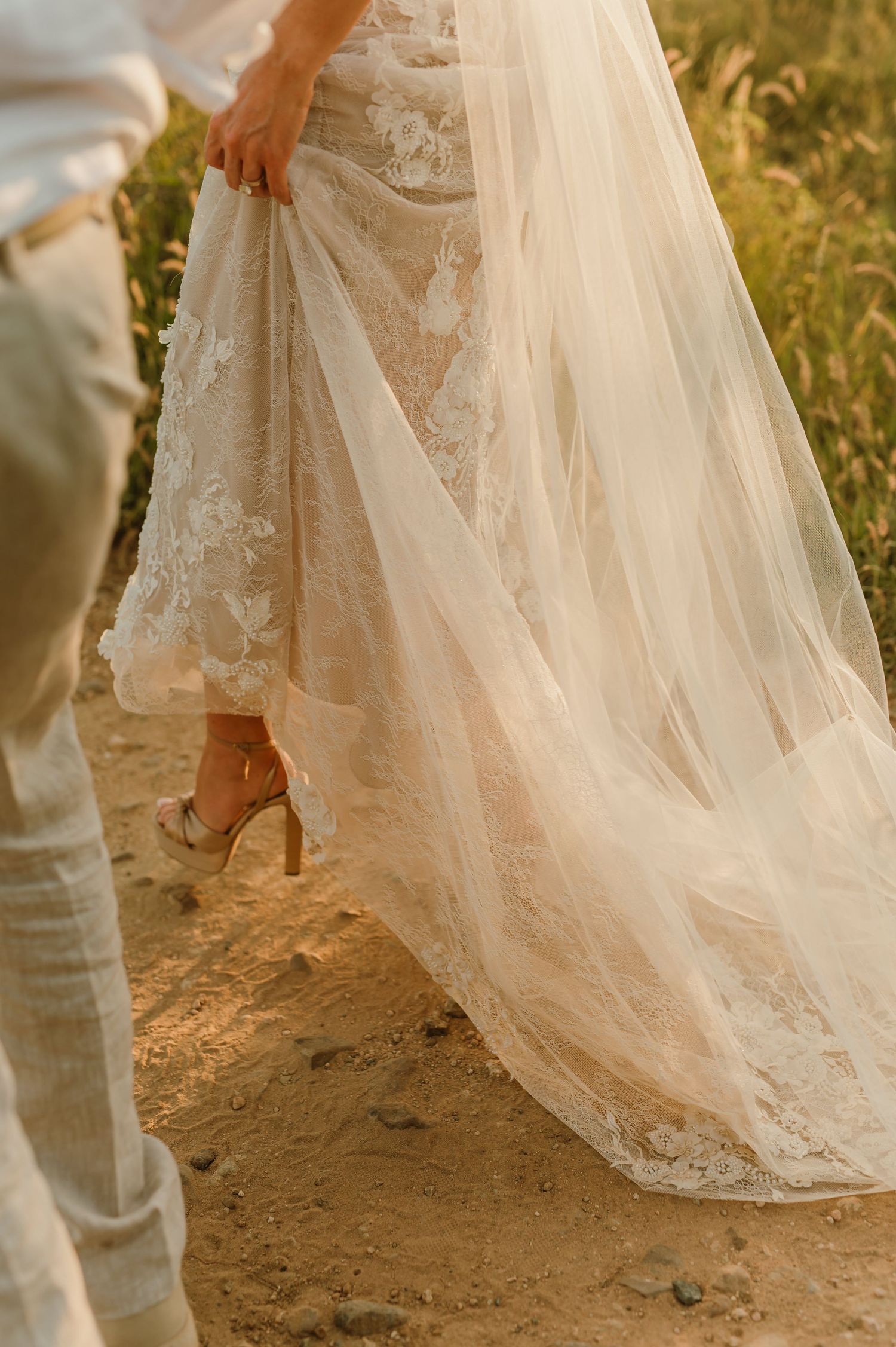 Close-up of bride in lace wedding gown with tulle train walking on sandy path in golden light.