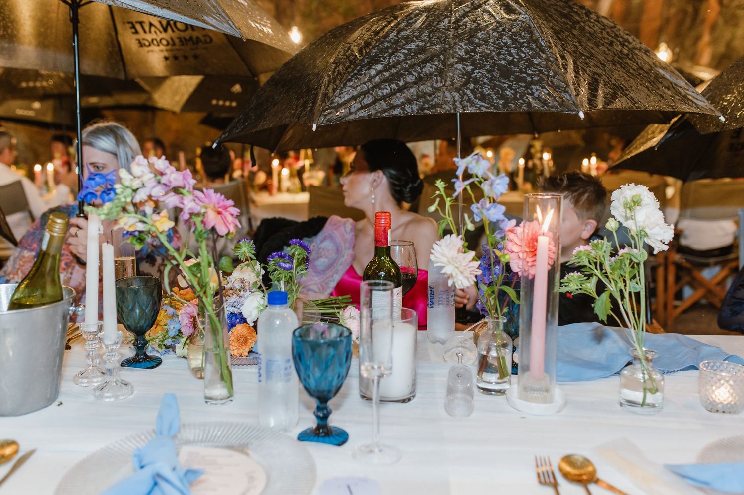 Colorful wildflower centerpieces with candles and blue glassware on white wedding reception table.
