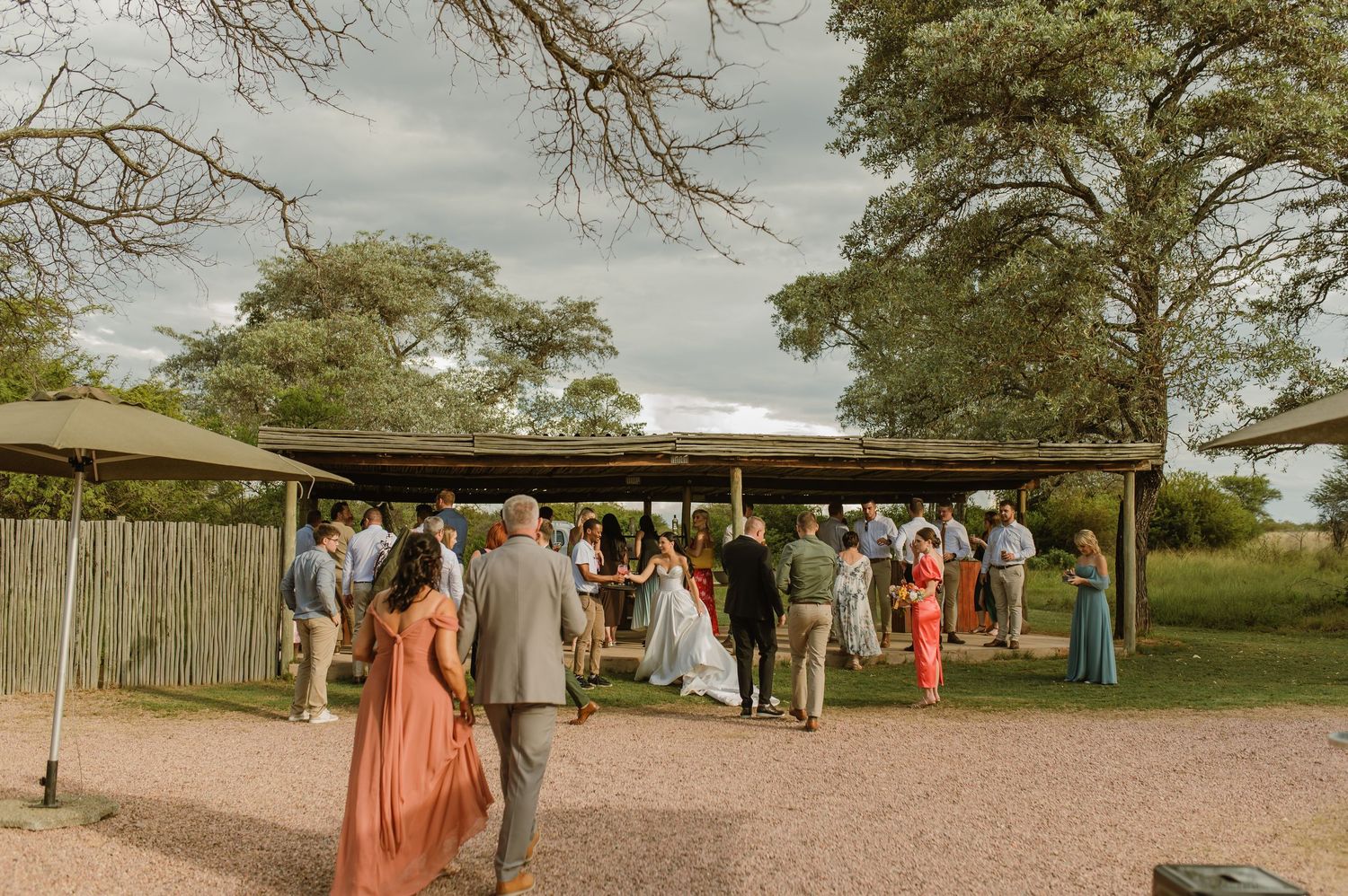 Wedding guests mingling under a shaded pavilion surrounded by trees at an outdoor venue.