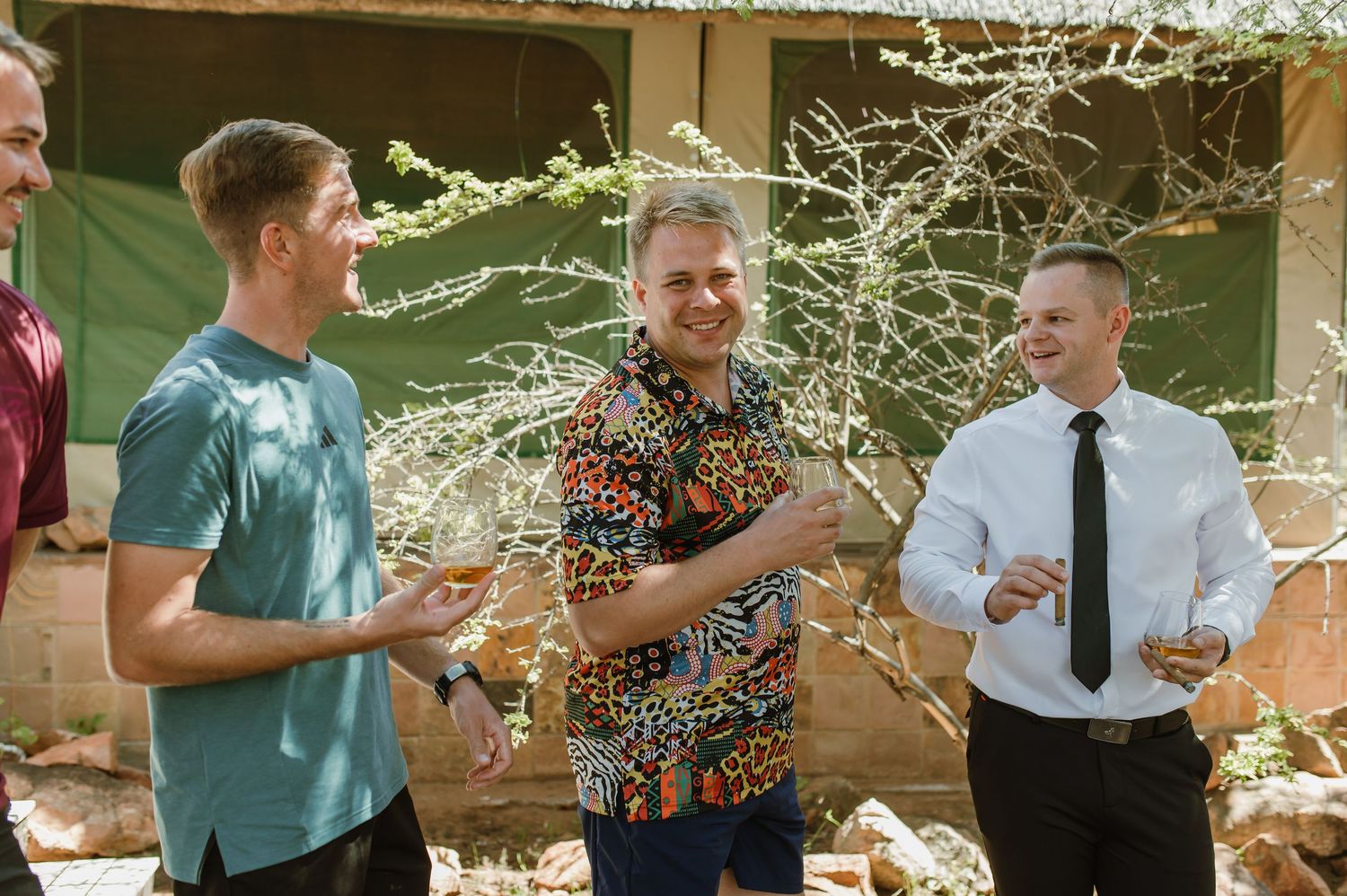 Four men laughing and socializing outdoors in casual and formal attire at a sunny wedding celebration.