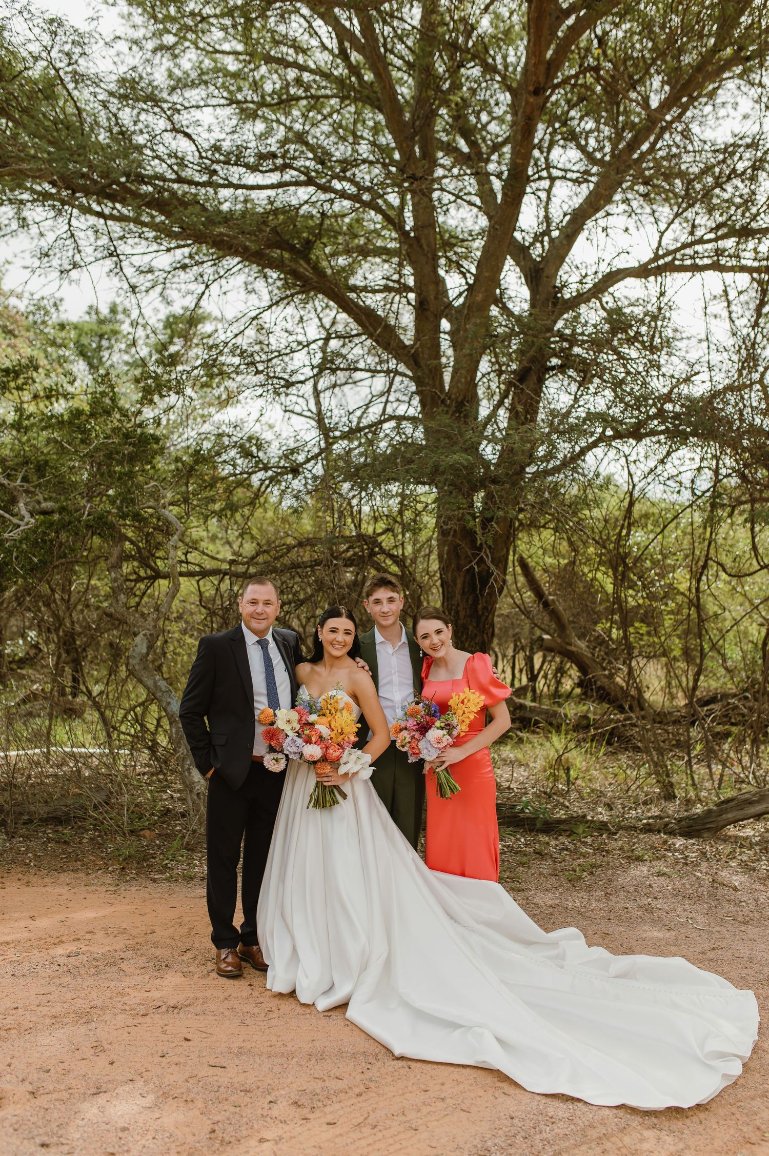 Wedding party of four poses under a tree with colorful bouquets on red dirt path.