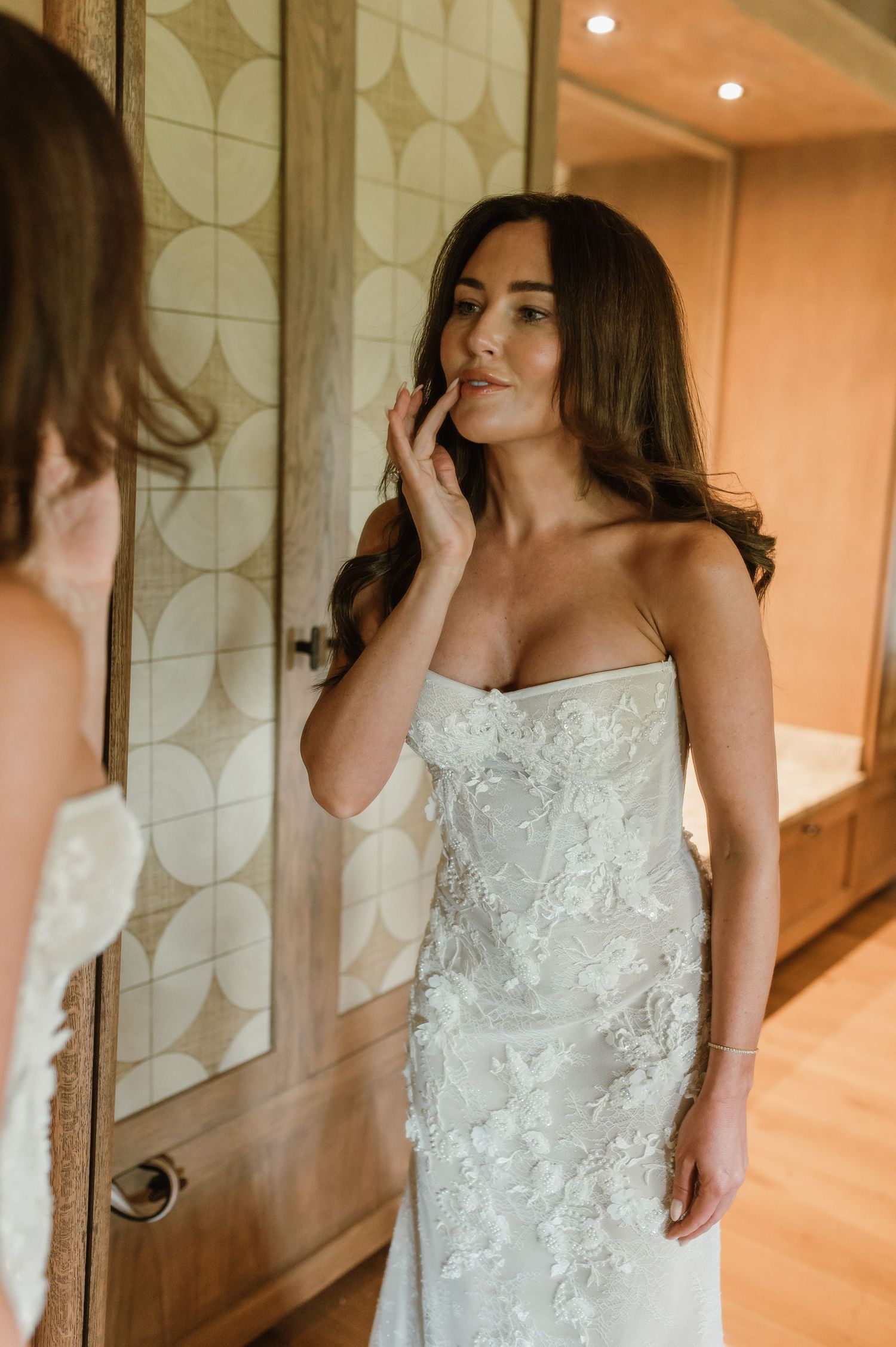 Bride in strapless lace wedding dress touching her lips while gazing into a mirror.