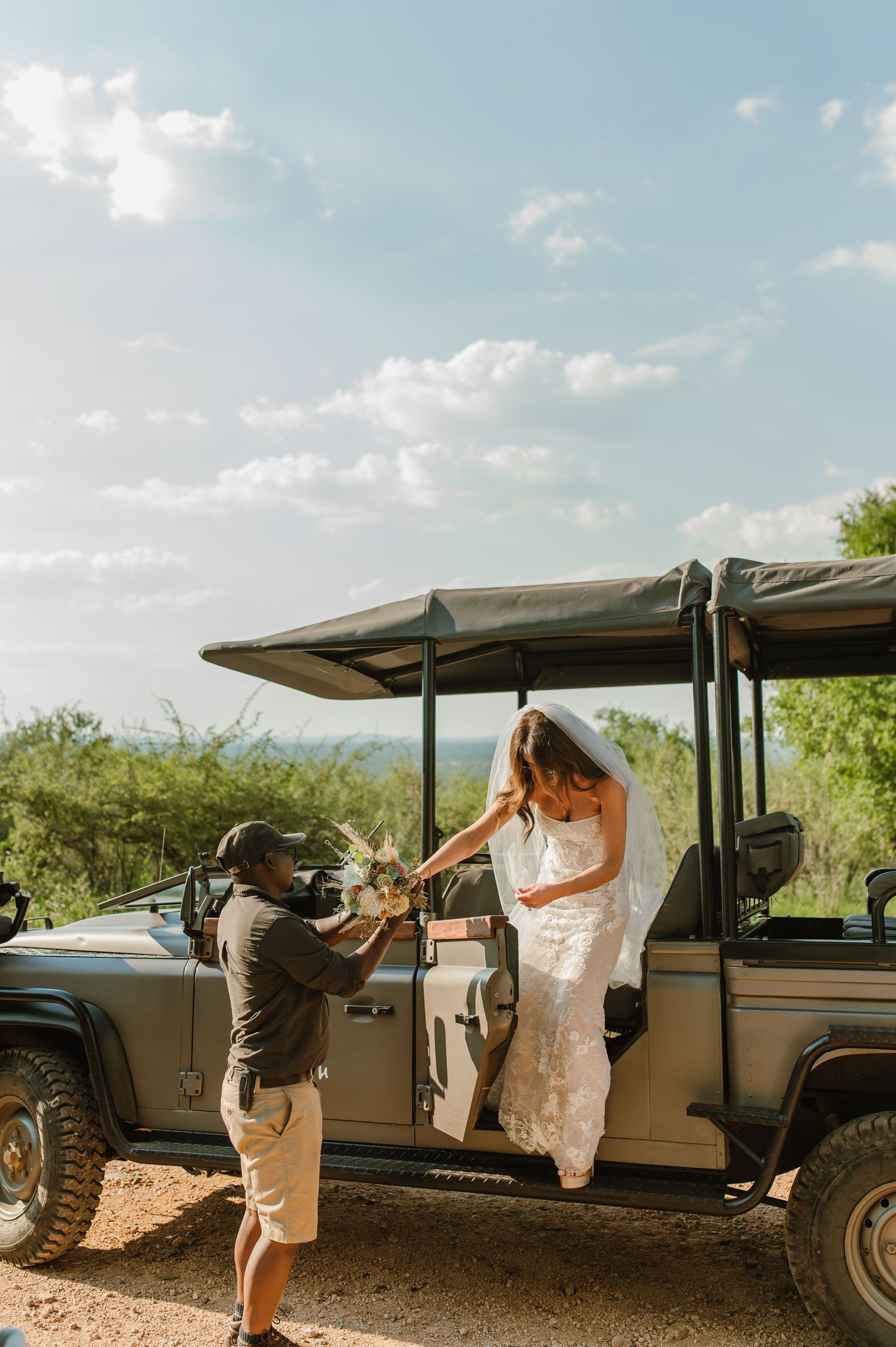 Bride in lace wedding gown being assisted from safari Land Rover by guide on sunny African bush day.
