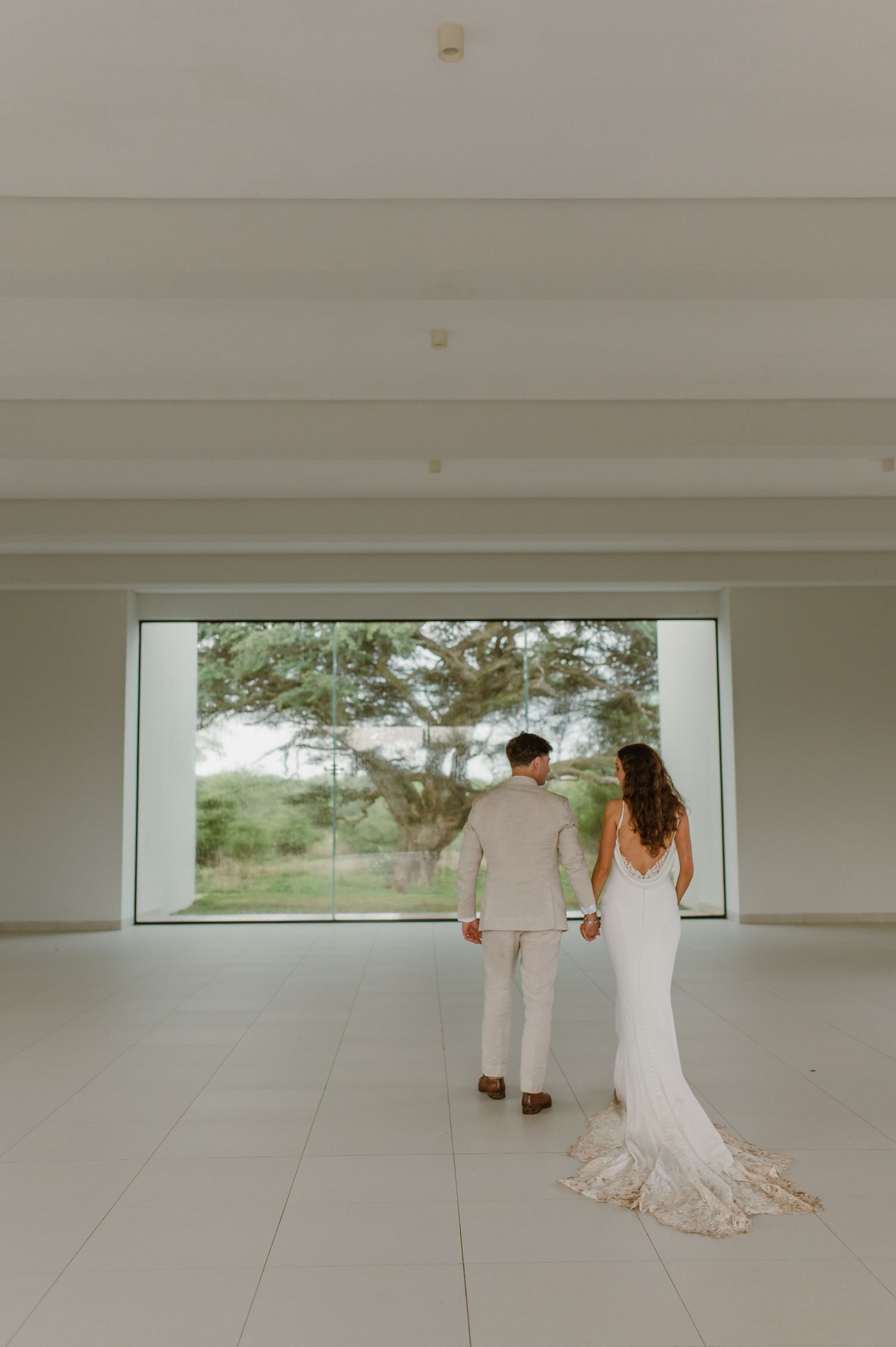 A wedding couple stands before a large window in a modern white interior space.