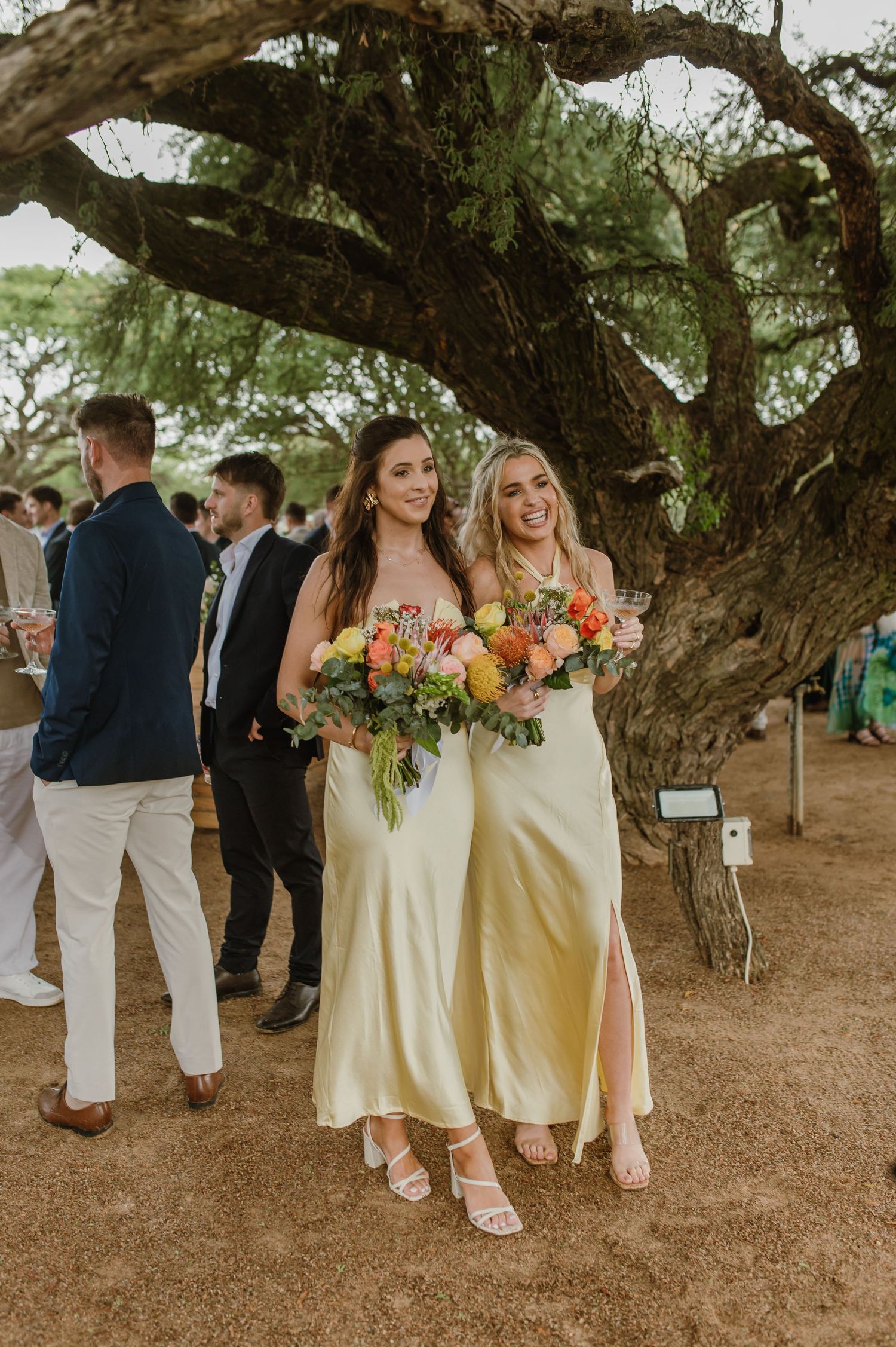 Bridesmaids in cream dresses carry bouquets under large tree at outdoor wedding.