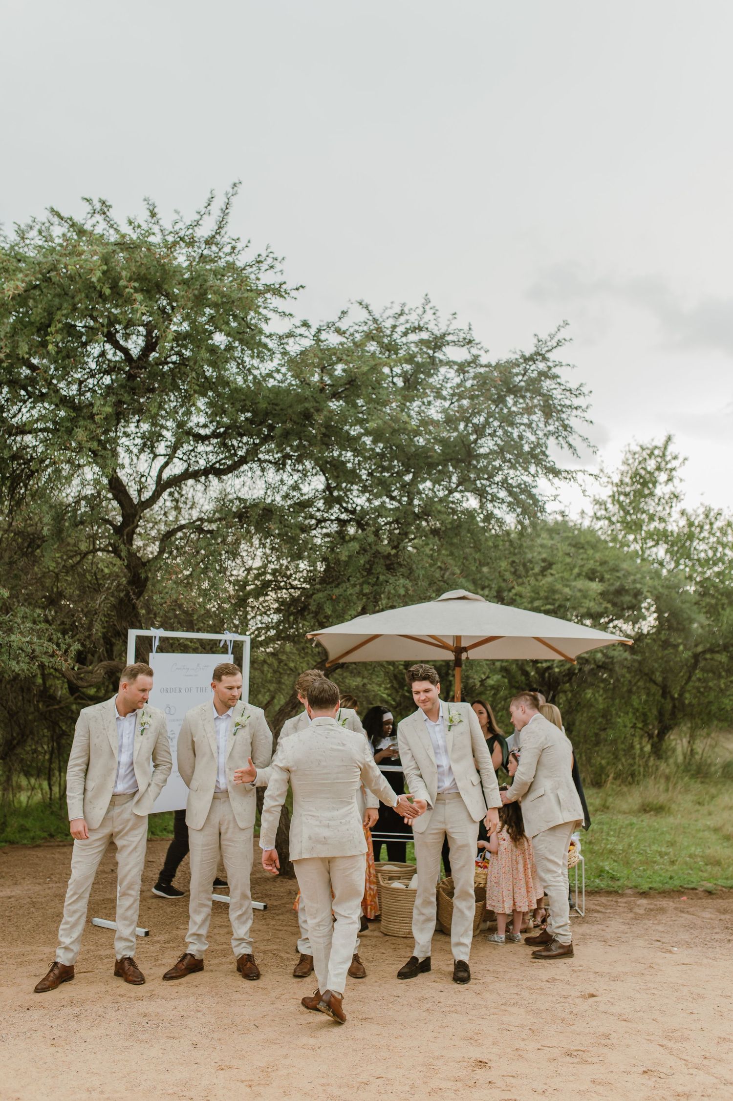Groomsmen in light beige suits gathering outdoors under trees at a wedding venue.