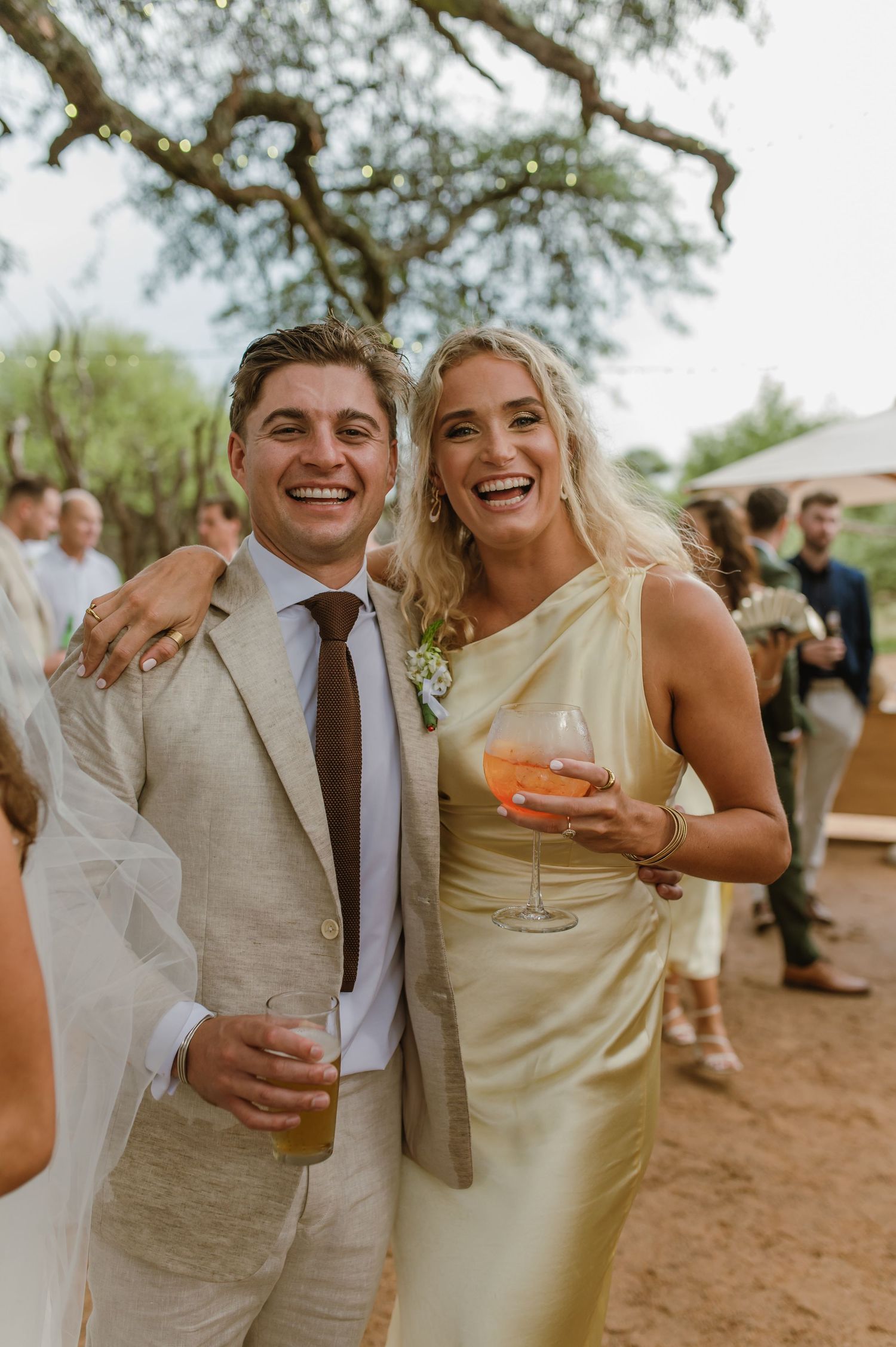 Wedding party members in light colored attire laugh and celebrate with drinks outdoors.