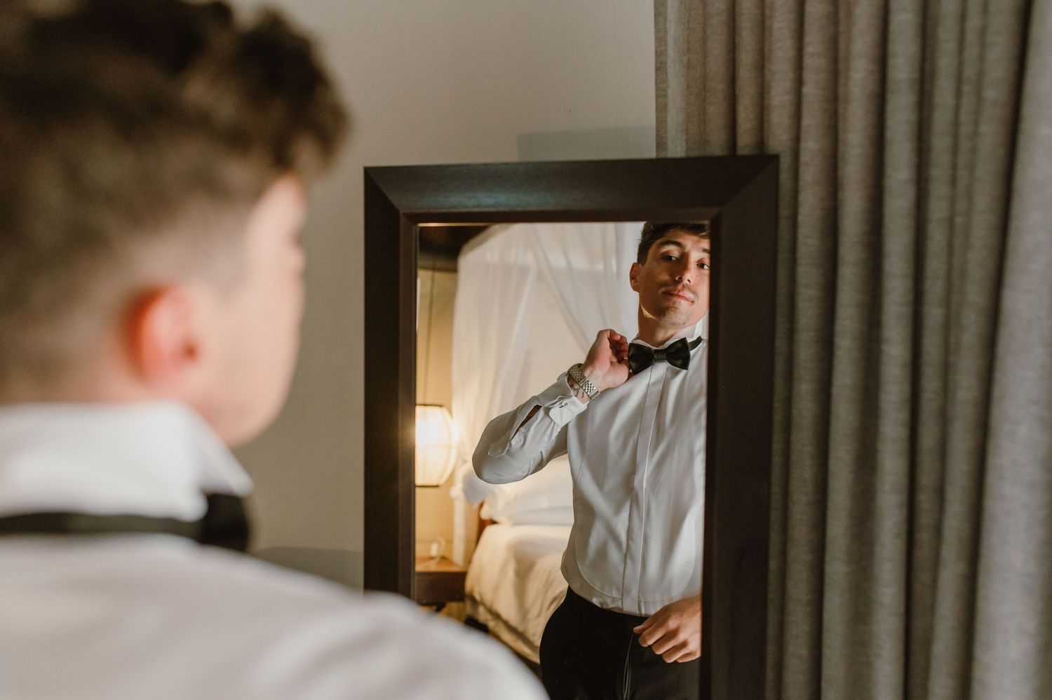 A person adjusting their tie while looking in a mirror with soft lighting and curtains in the background.
