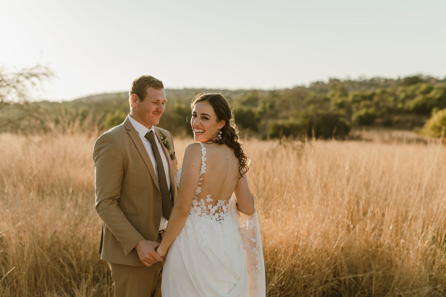 A romantic sunset wedding photo of a couple in a golden field wearing a tan suit and lace back wedding dress.